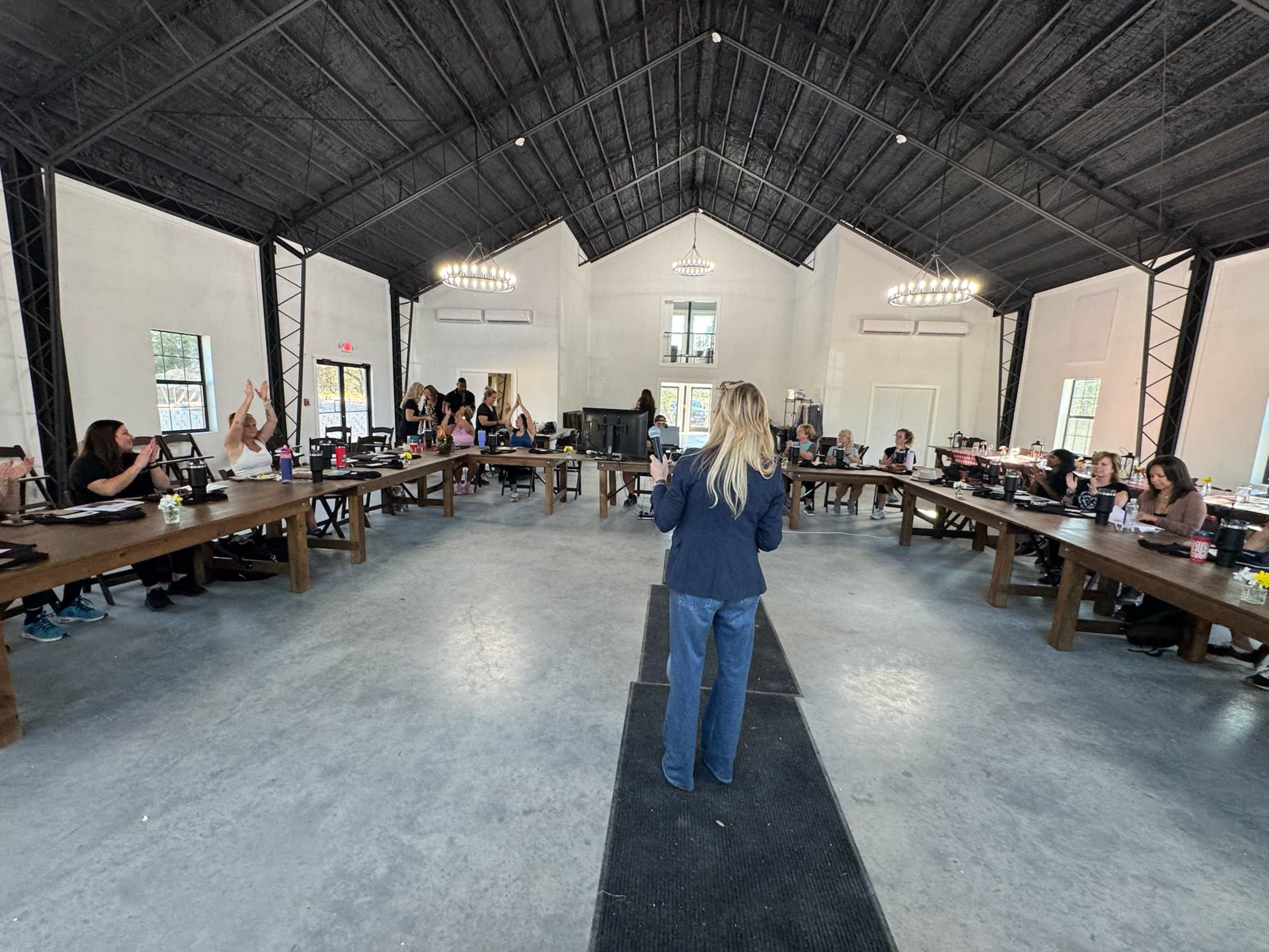 A group of people is seated around large wooden tables in a spacious room, with some individuals raising their hands as a woman stands at the front speaking.