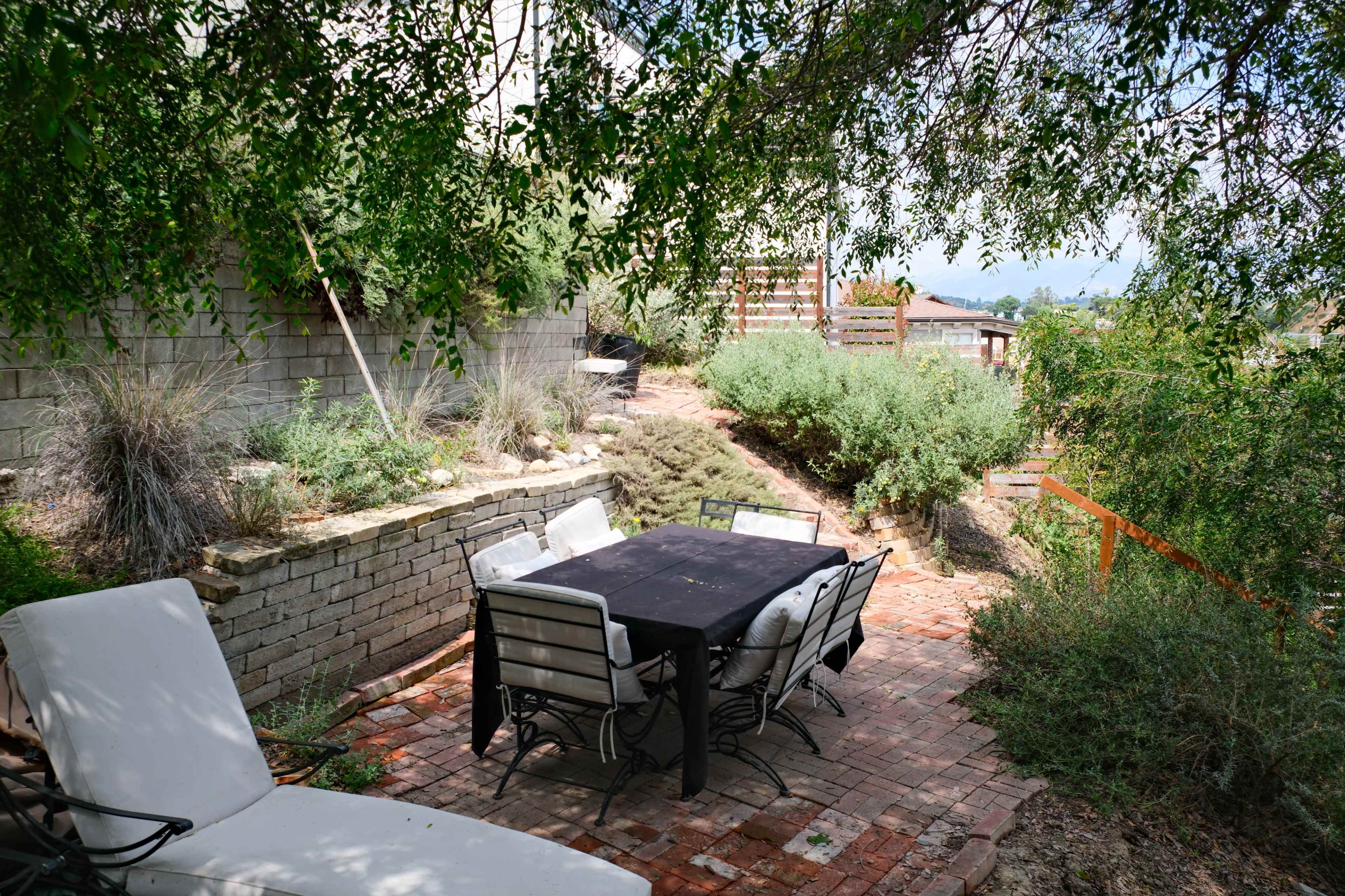 A shaded outdoor patio area features a black table surrounded by chairs, with a backdrop of greenery and a terraced landscape.