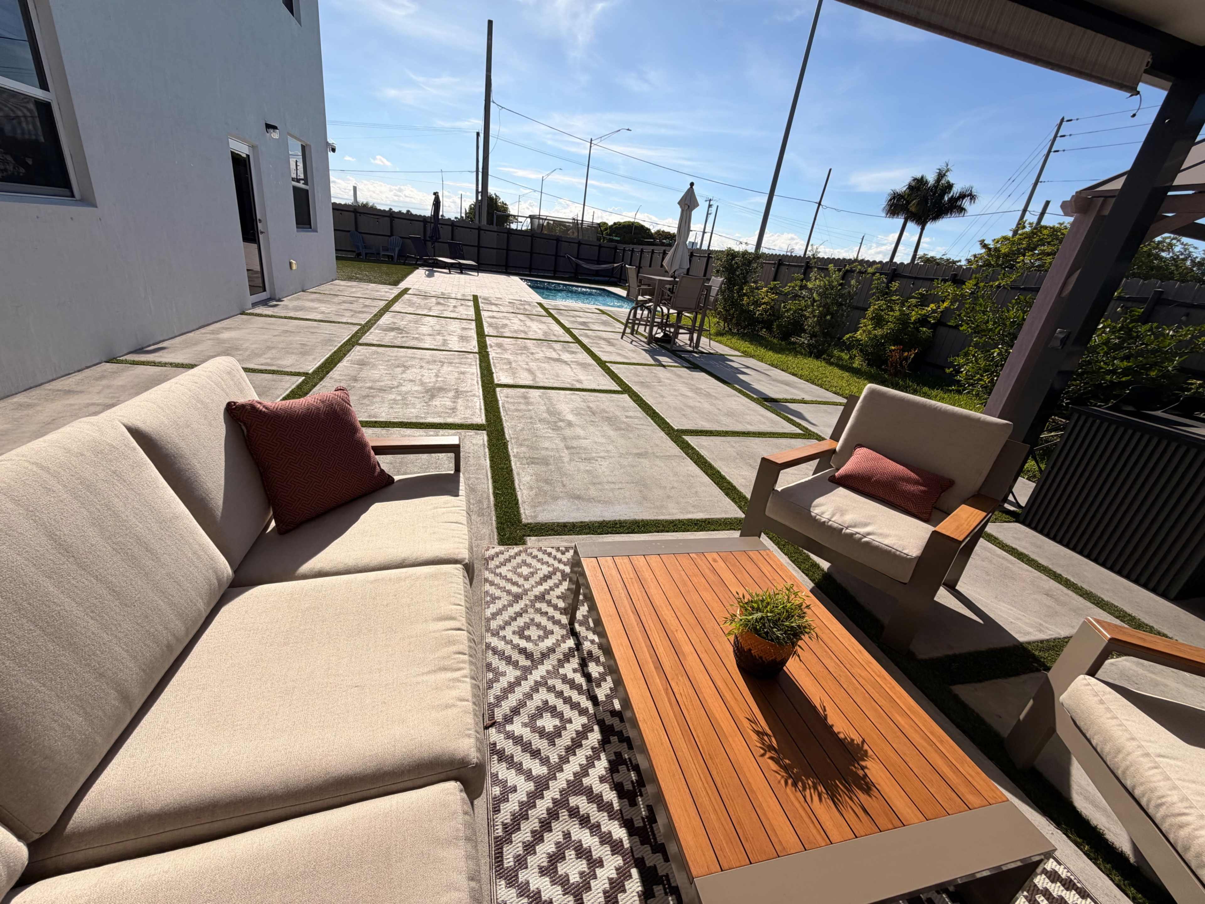 The image shows a patio area with outdoor furniture, a swimming pool in the background, and a clear blue sky.