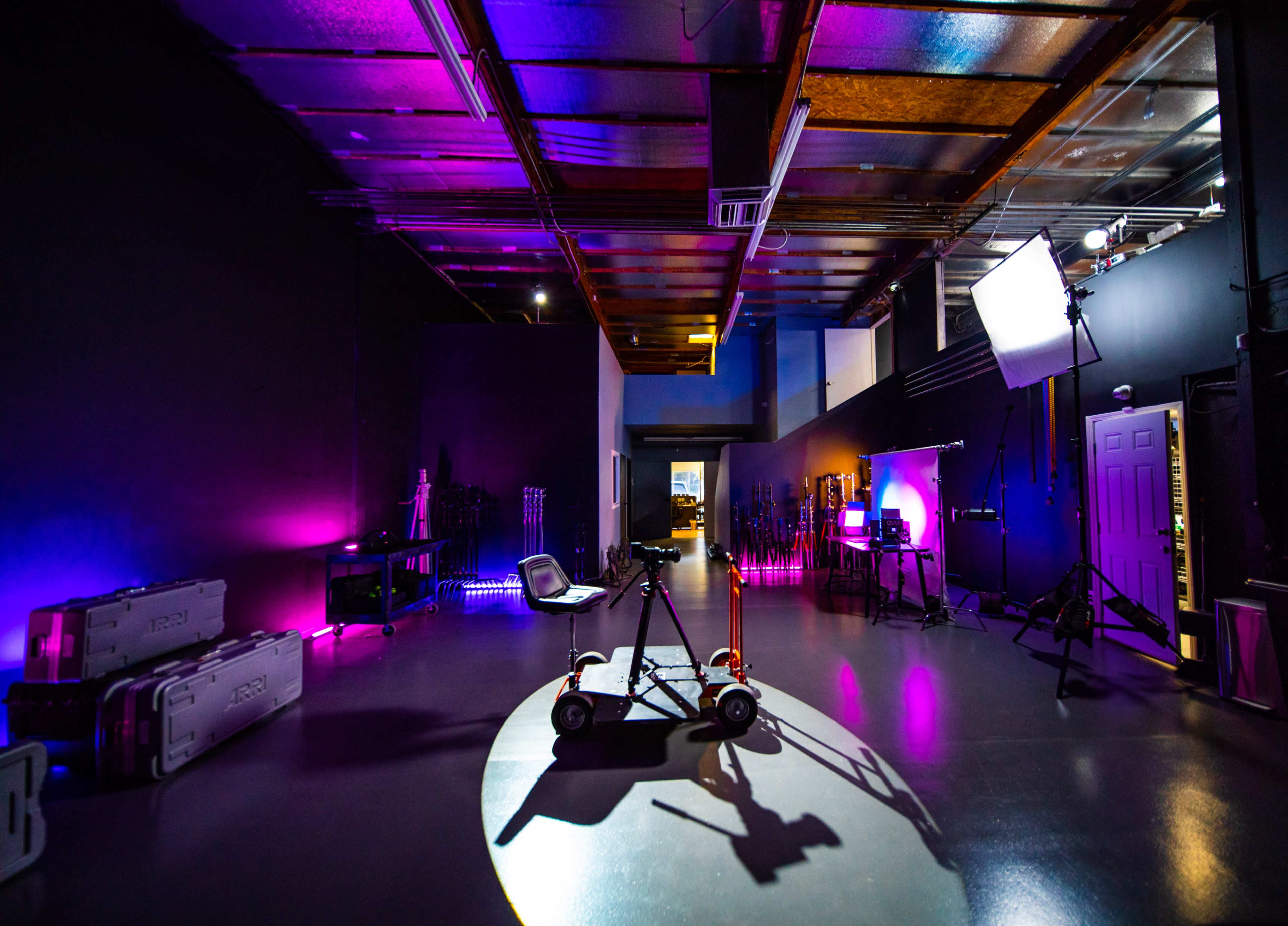 A spacious studio with purple and blue lighting, featuring camera equipment, a light setup, and a wheeled cart in the foreground.