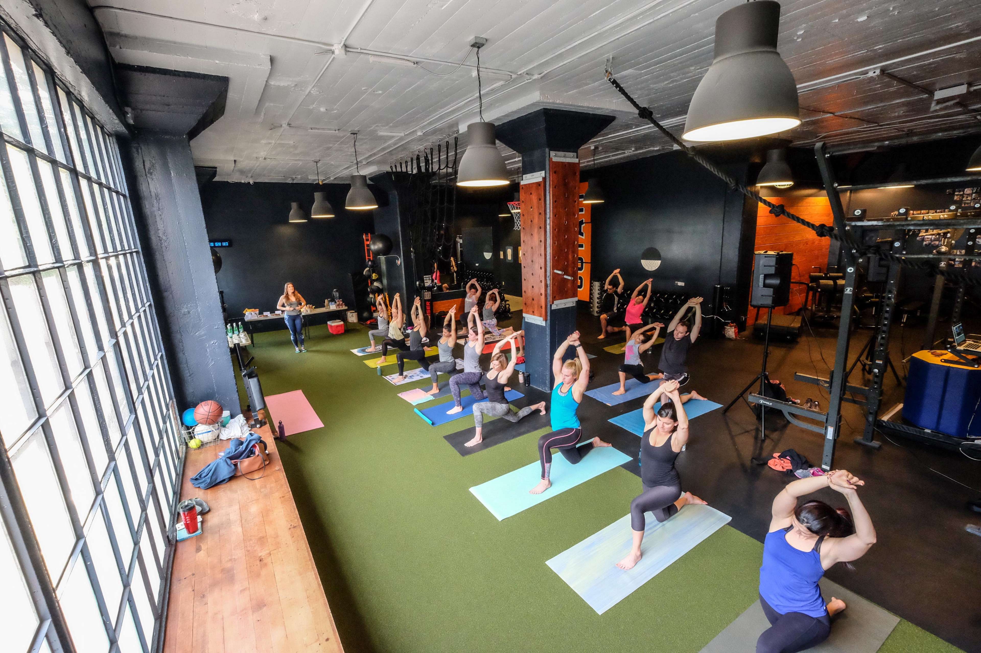 A group of people is practicing yoga on mats in a spacious gym with large windows and various fitness equipment in the background.