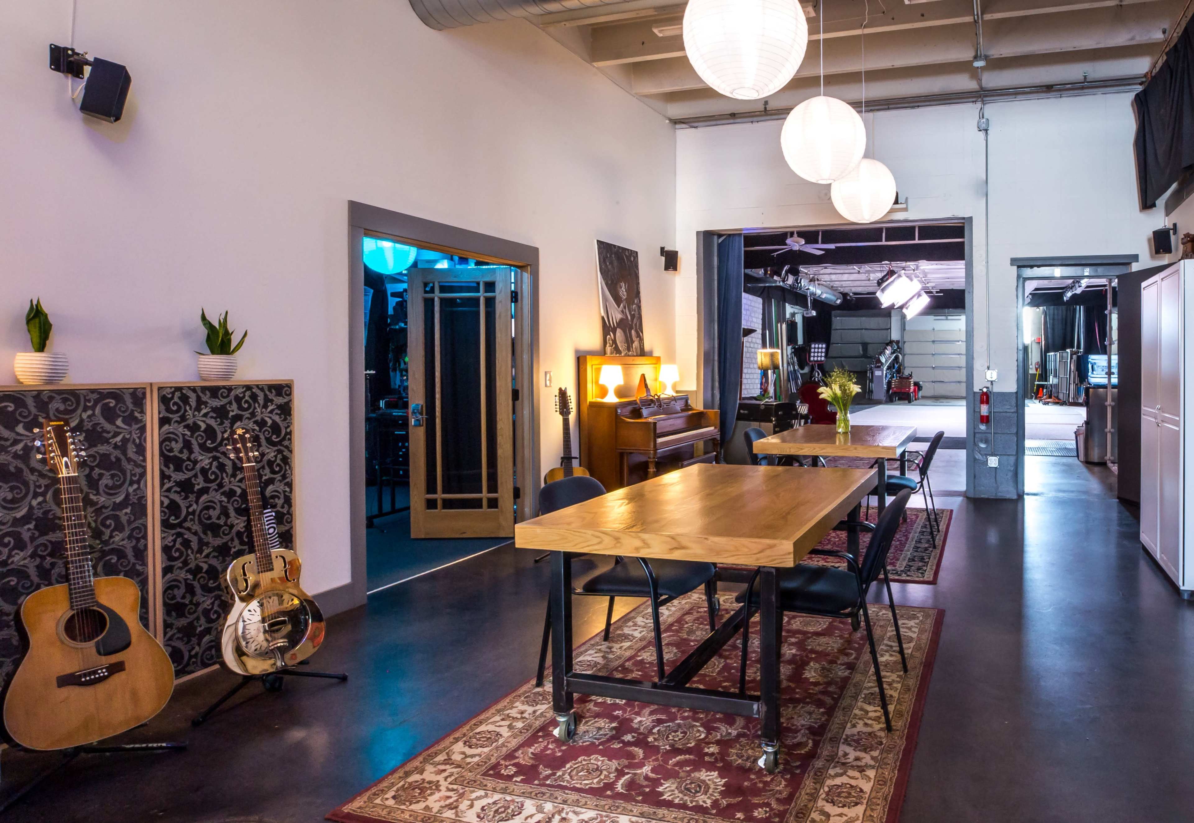 A spacious studio-like interior with a wooden table, guitars leaning against the wall, and a series of round pendant lights hanging from the ceiling.