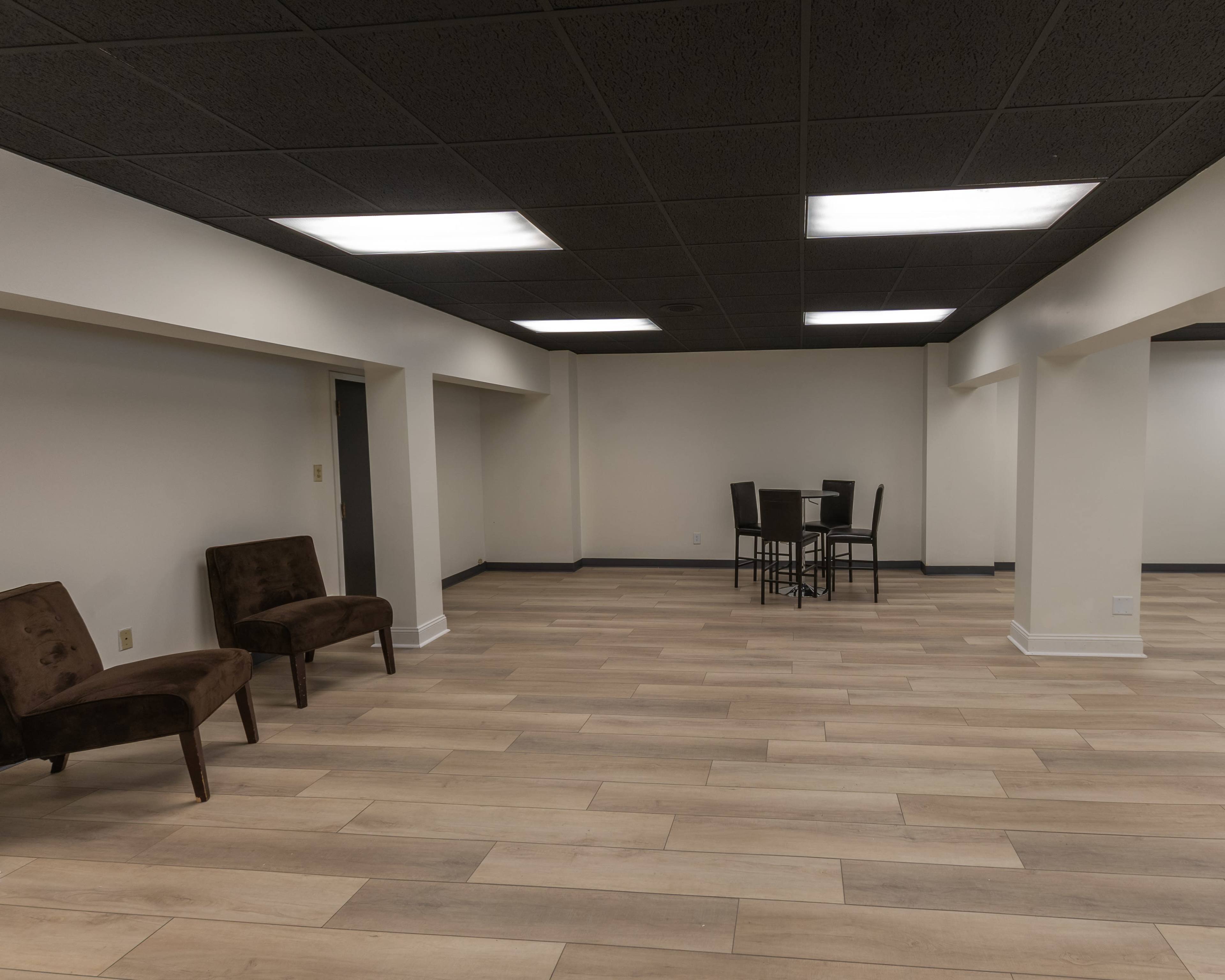An empty room with light-colored wooden flooring, beige walls, and a black ceiling, featuring two brown chairs and a small dining table with black chairs.