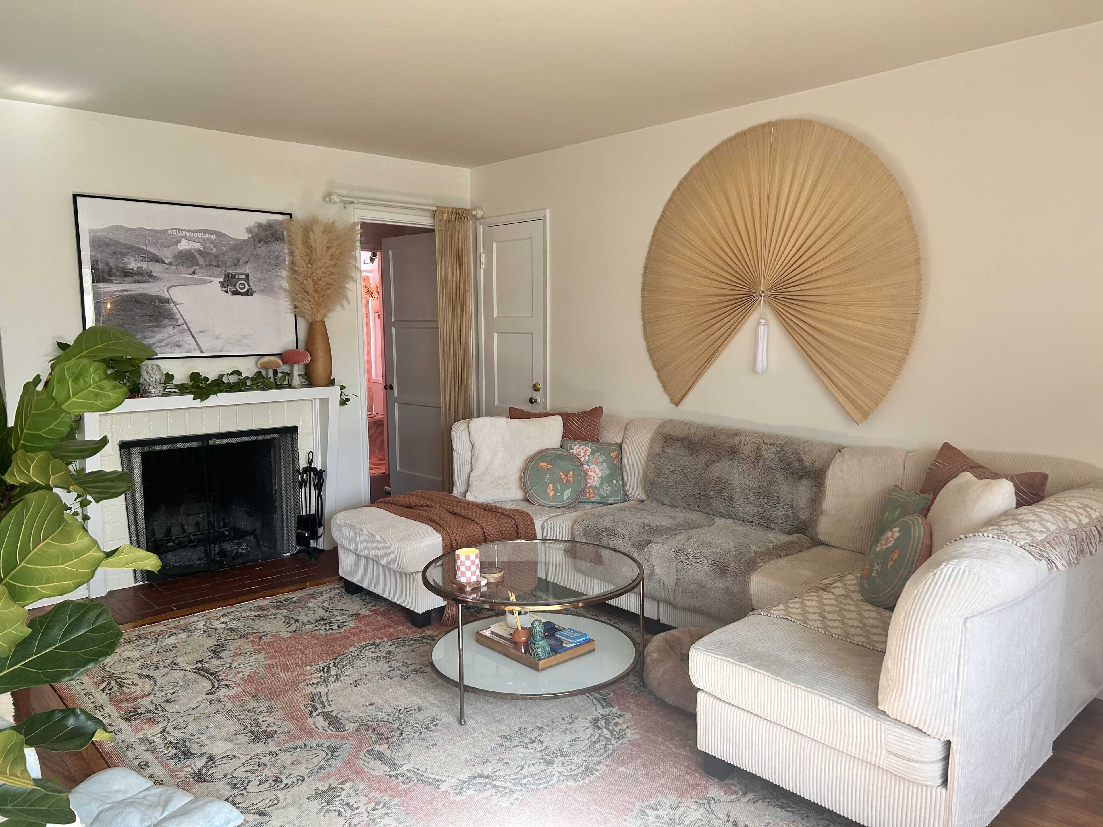 The living room features a large sectional sofa, a round glass coffee table, and a decorative wall hanging, with a fireplace and a doorway leading to another room.