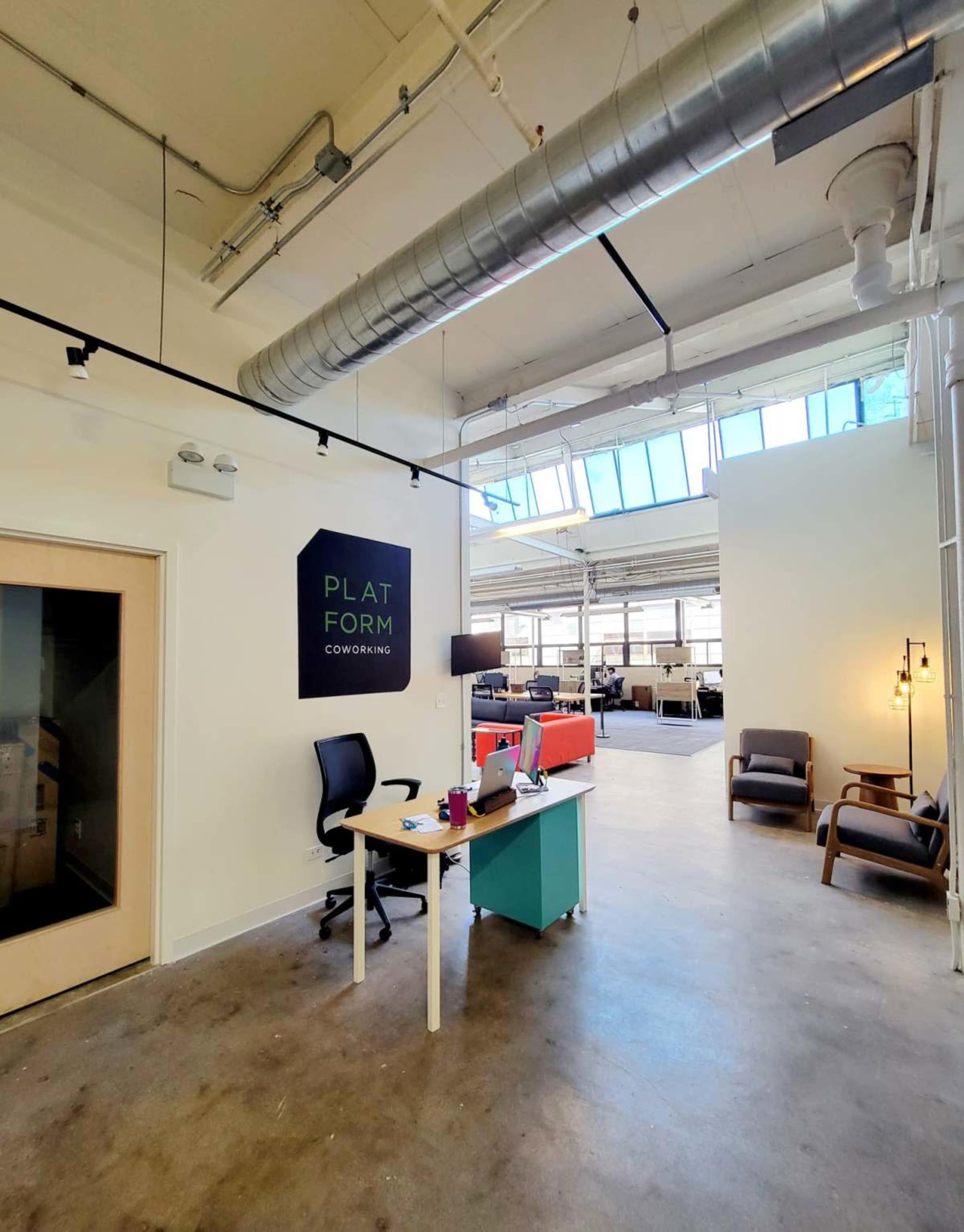 The image shows a modern coworking space with a reception desk, chairs, and large windows letting in natural light.