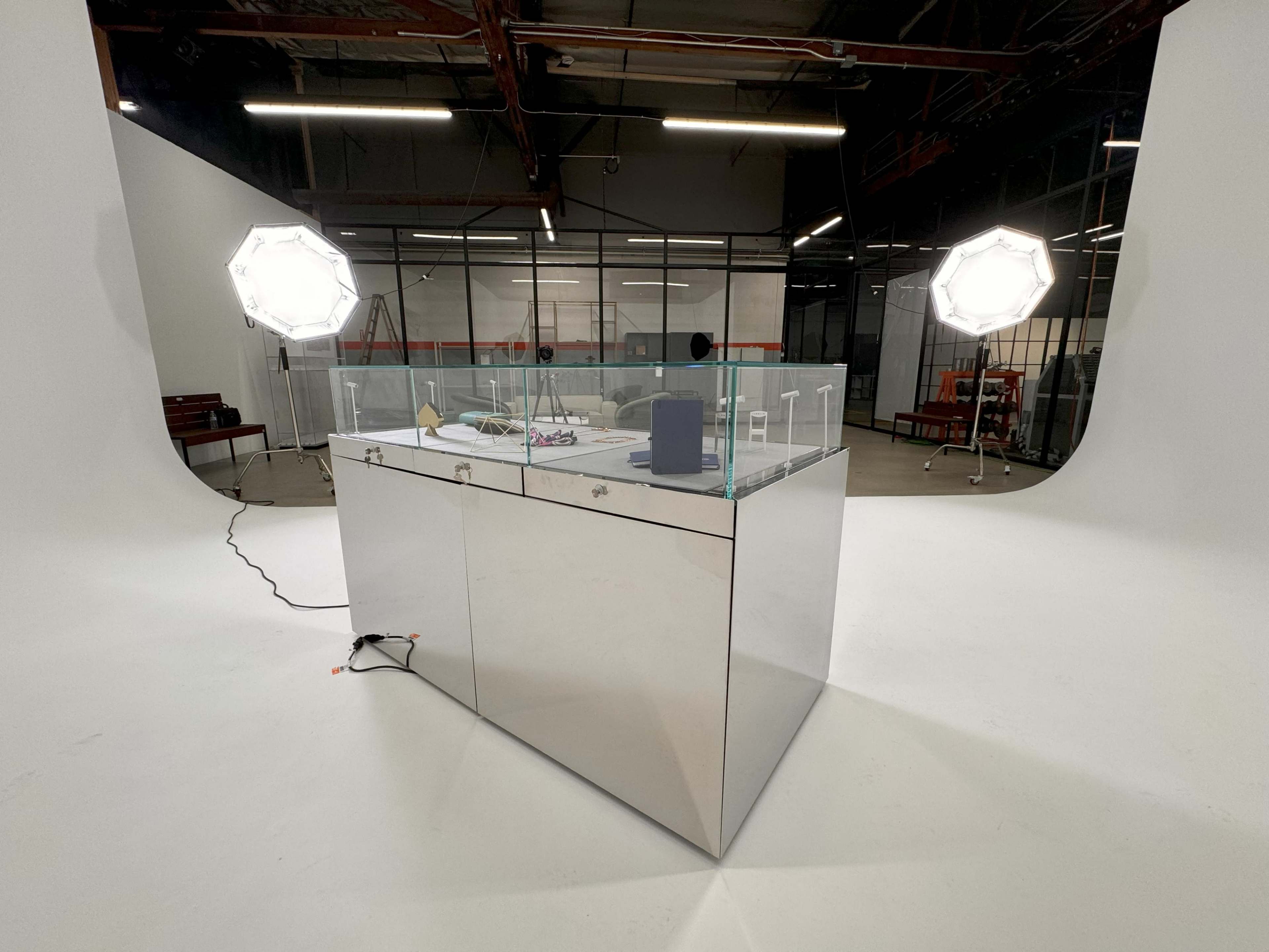 A reflective glass display case is positioned in the center of a well-lit studio with softbox lighting on either side.
