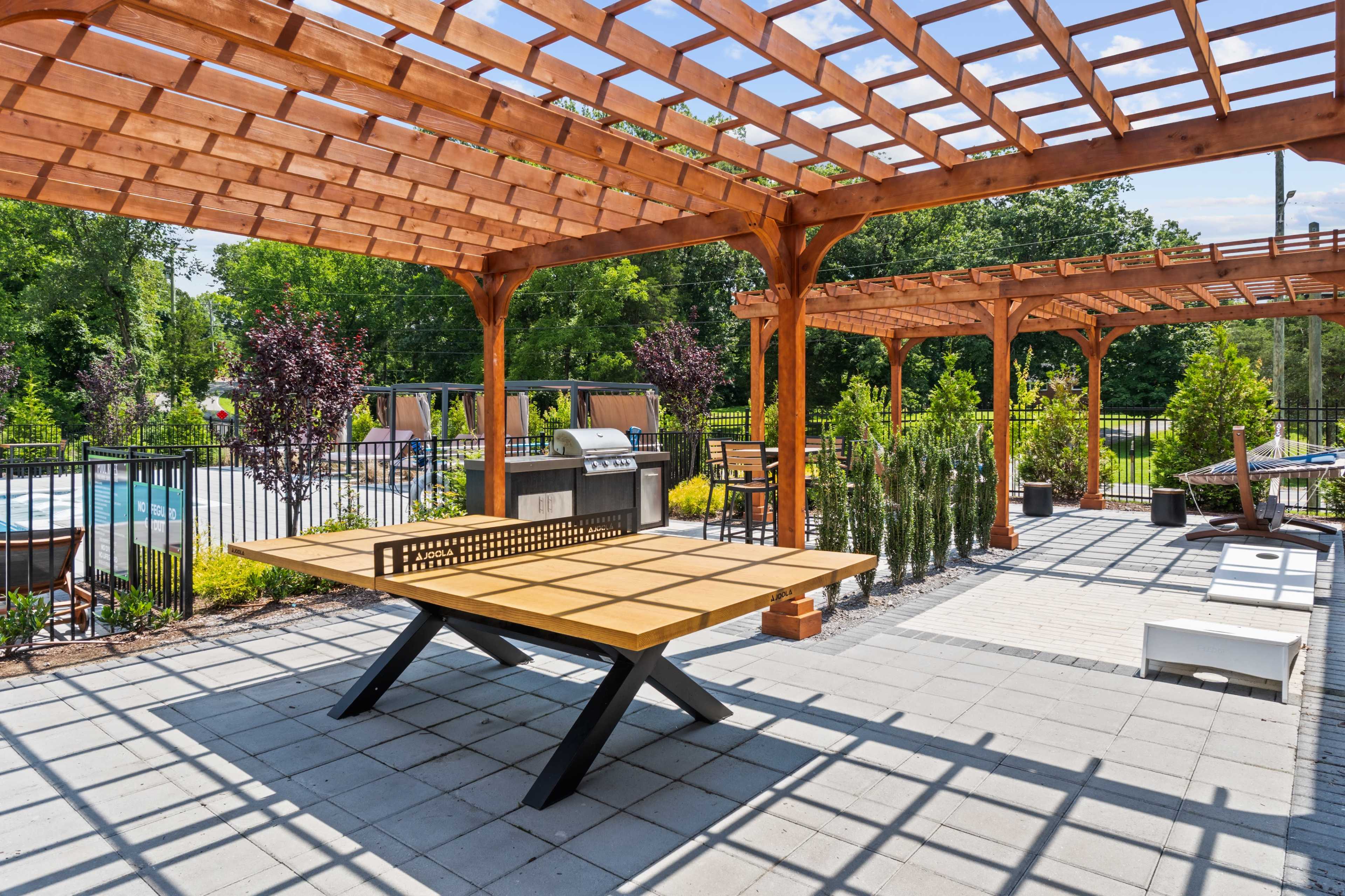 The image shows a landscaped outdoor patio area featuring a large wooden table, a barbecue grill, and pergolas with seating, surrounded by greenery and a swimming pool in the background.