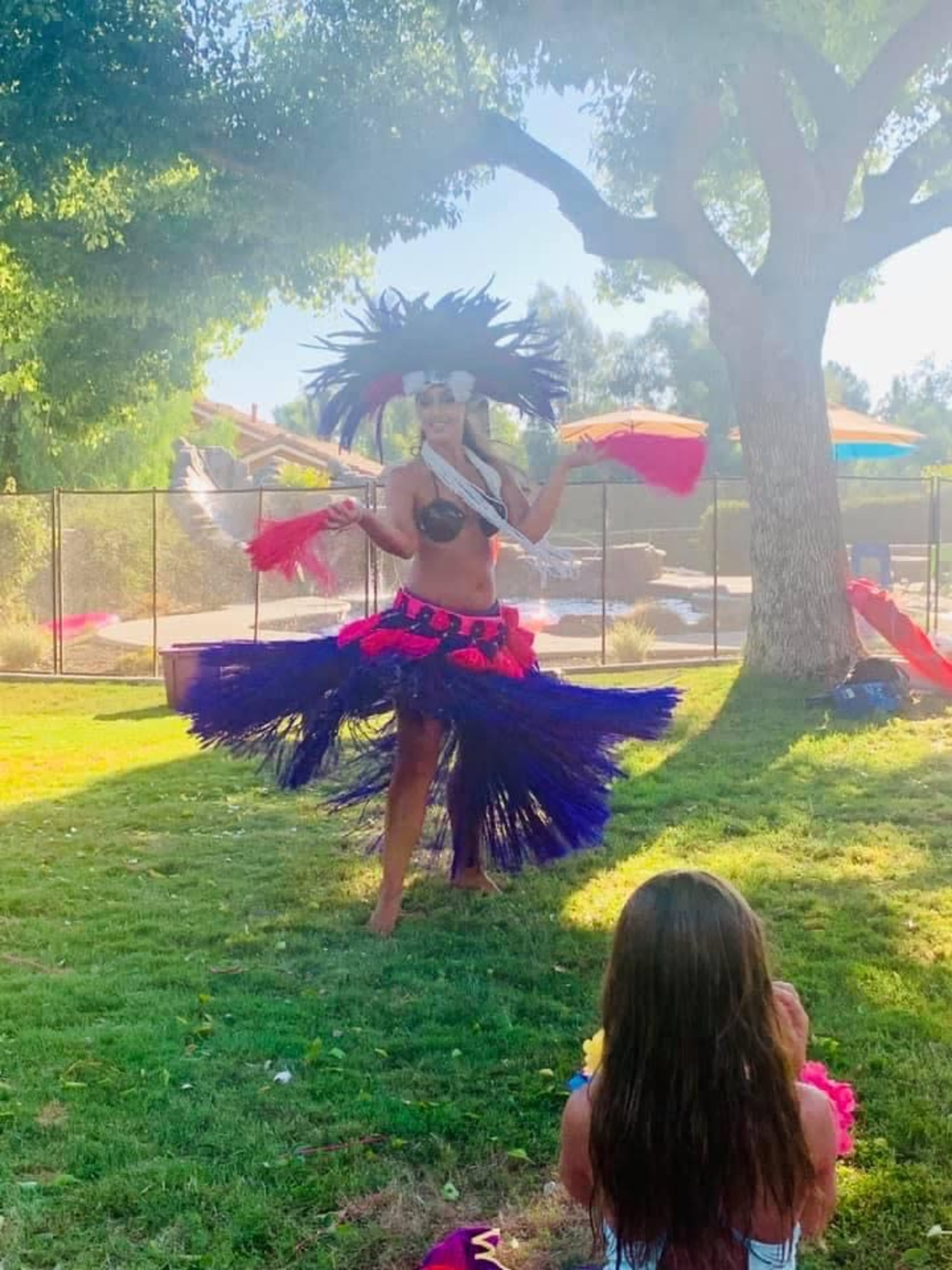 A performer in a colorful hula costume dances on a grassy area while a young girl watches from the foreground.