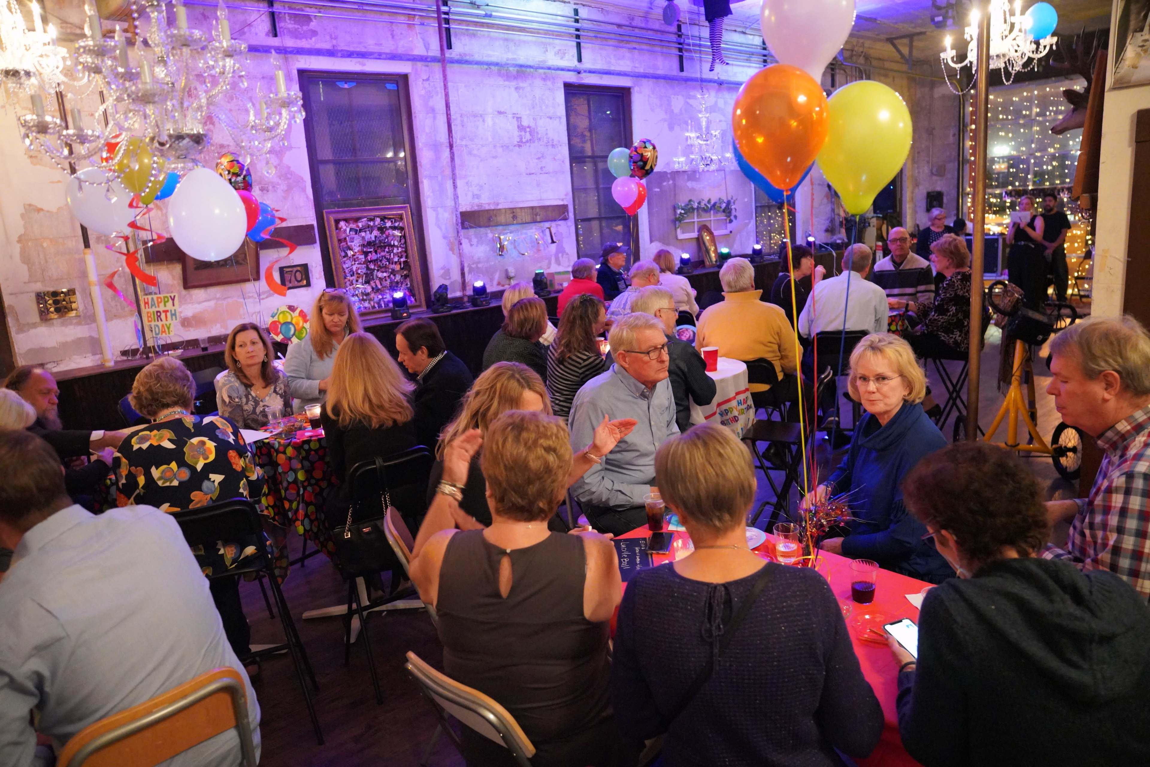A festive gathering takes place in a brightly decorated venue with tables of people socializing and colorful balloons in the air.