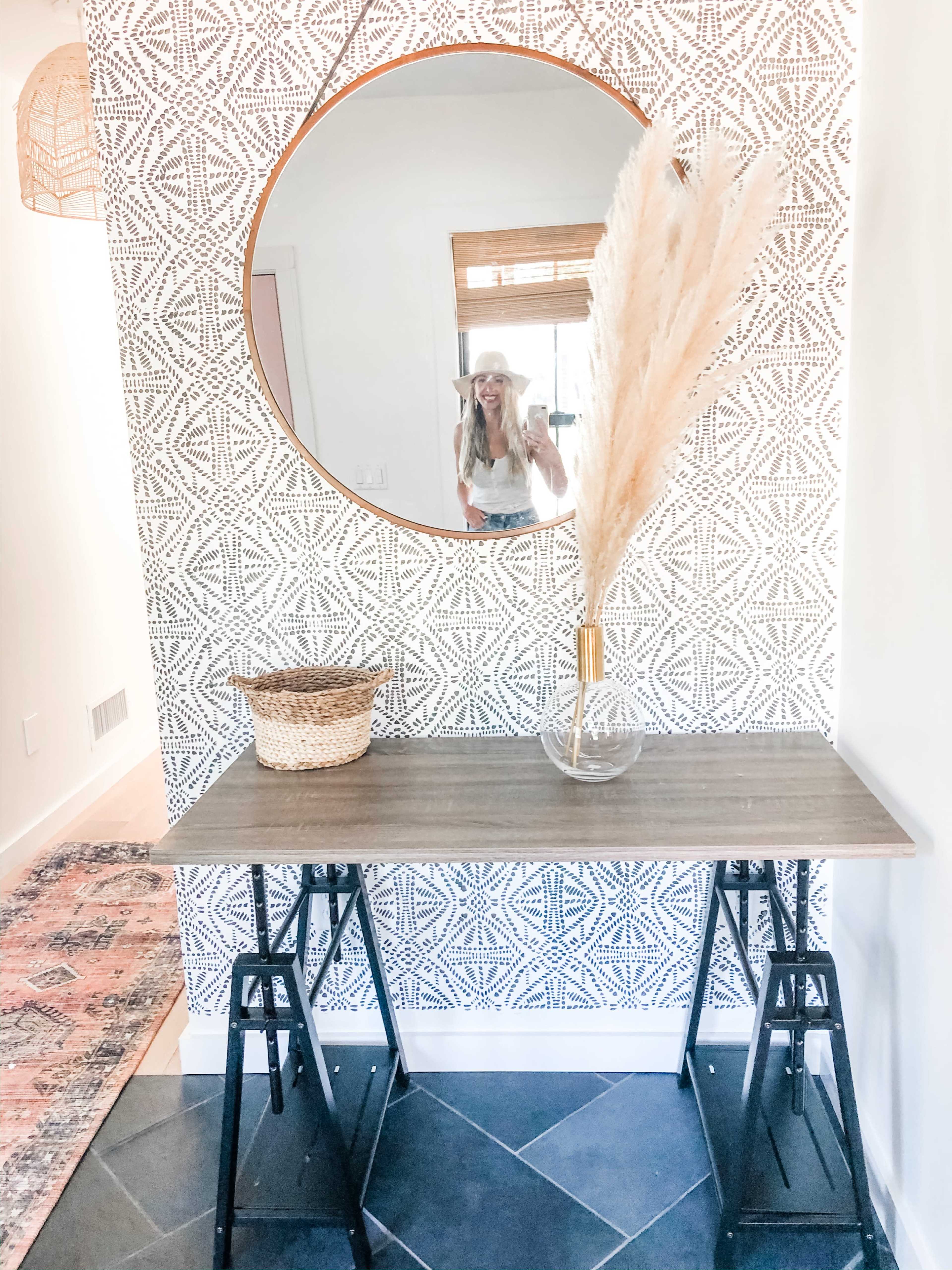 A mirrored round wall adorns a patterned backdrop, with a wooden table featuring a transparent vase and dried pampas grass, beside a woven basket.