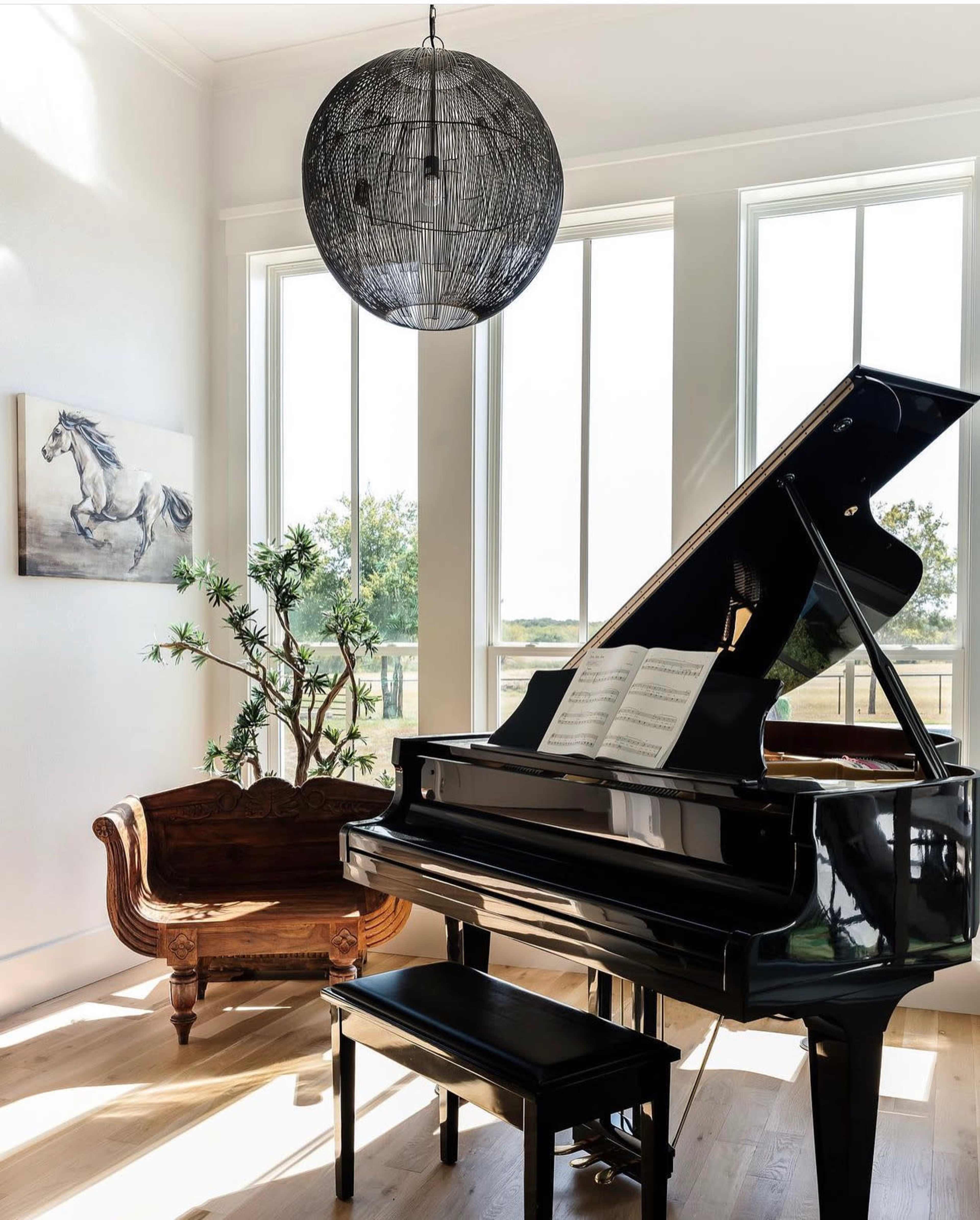 A grand piano with sheet music sits beside a vintage couch under a large, spherical light fixture in a bright room with large windows.