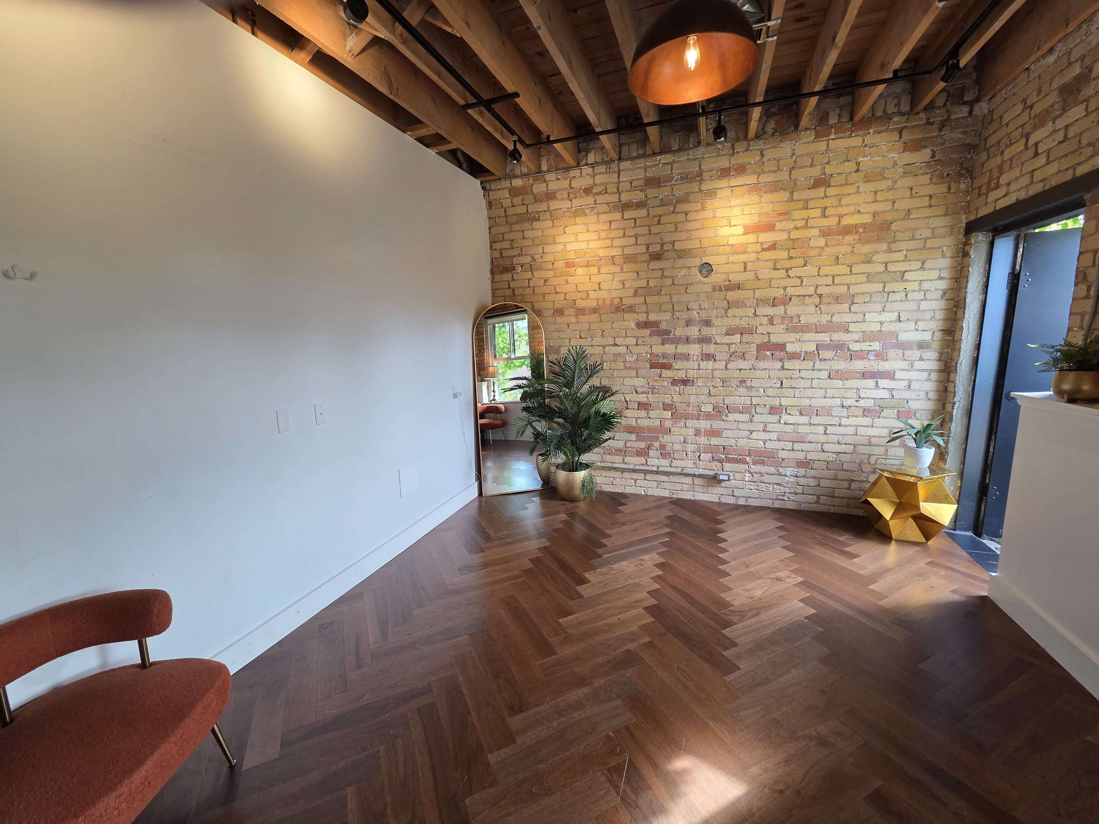 The room features a herringbone wooden floor, exposed brick walls, a mirror, a potted plant, and a decorative gold object.