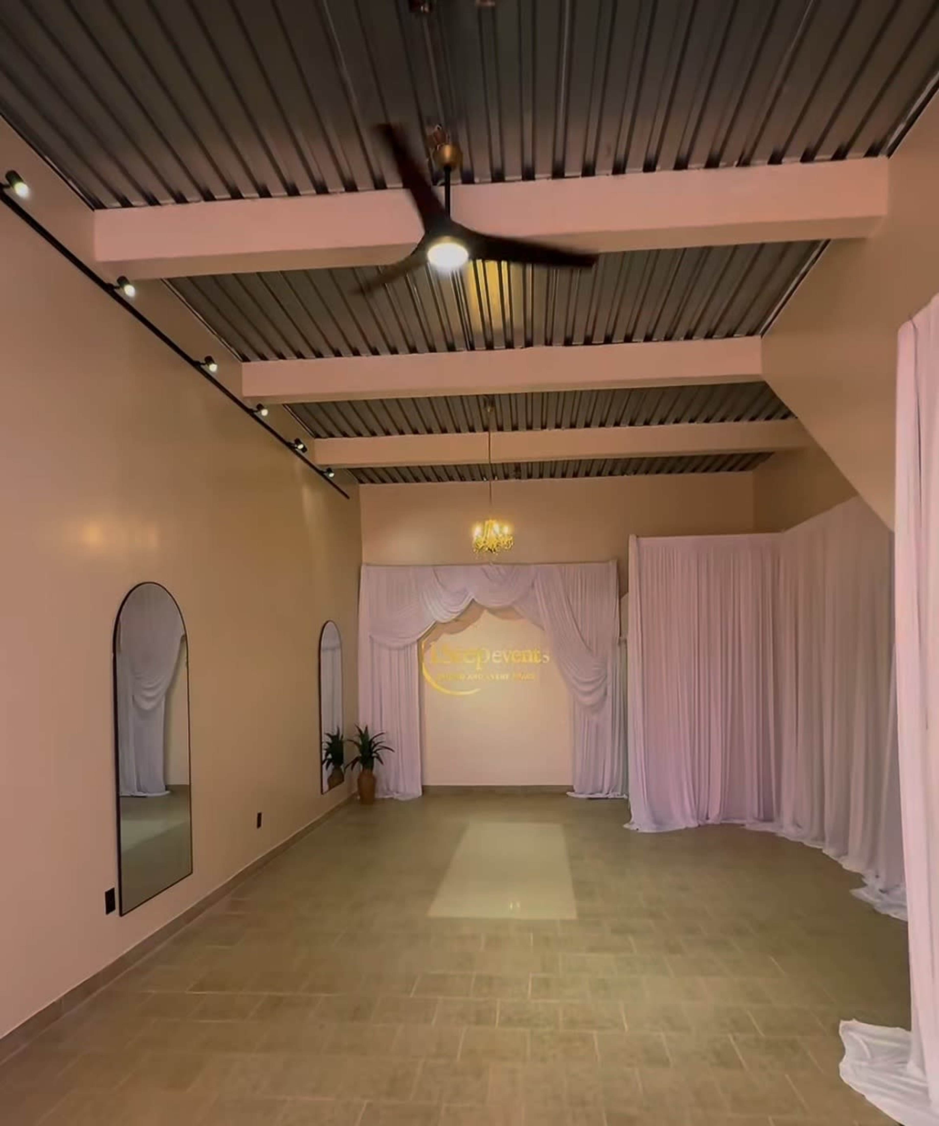 The image shows a spacious room with beige walls, a tiled floor, and draped white fabric along one side, featuring a mirror and a decorative light fixture.