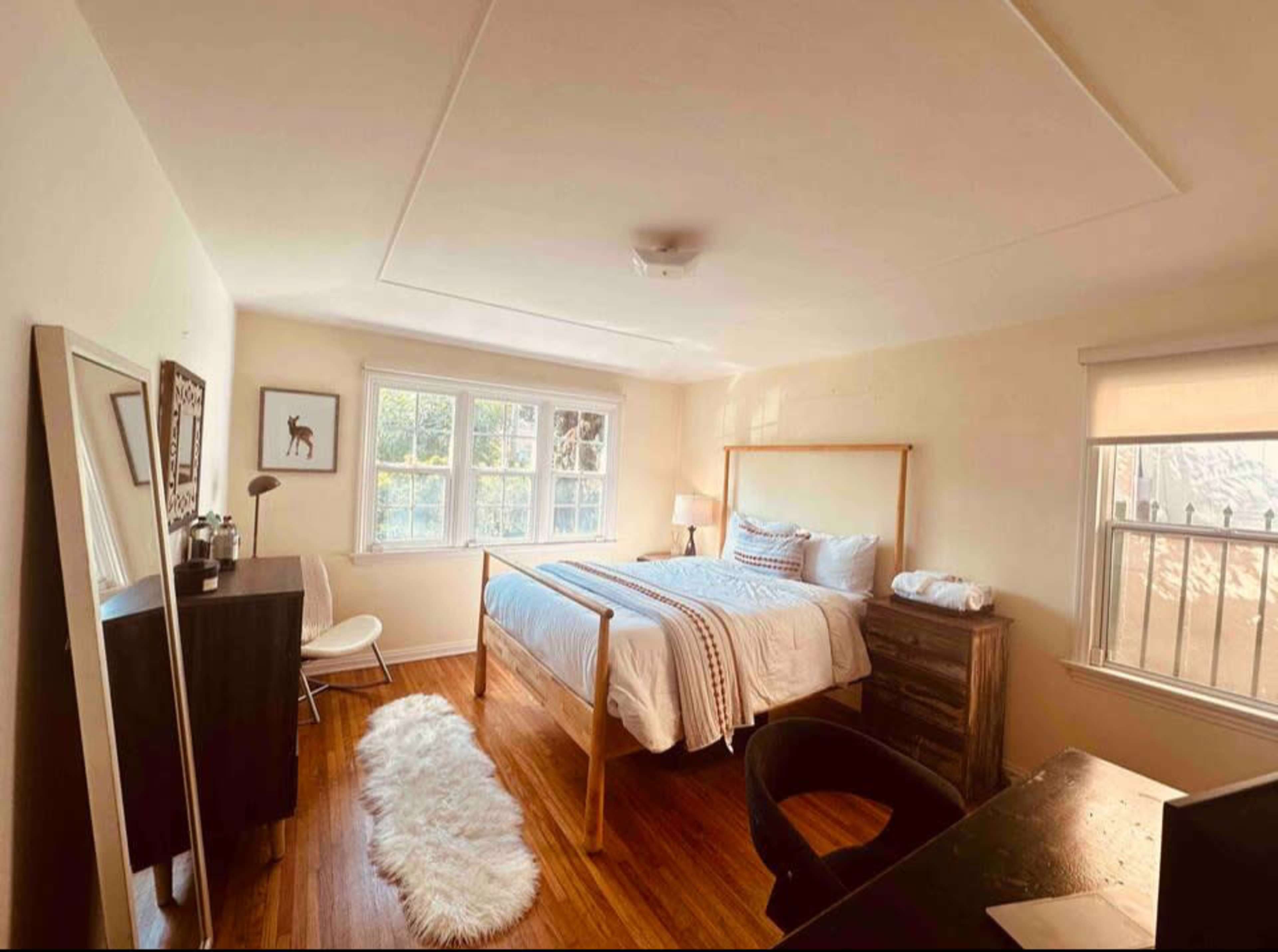 The image shows a well-lit bedroom featuring a bed with white bedding, a desk, a chair, a dresser, and a large mirror.