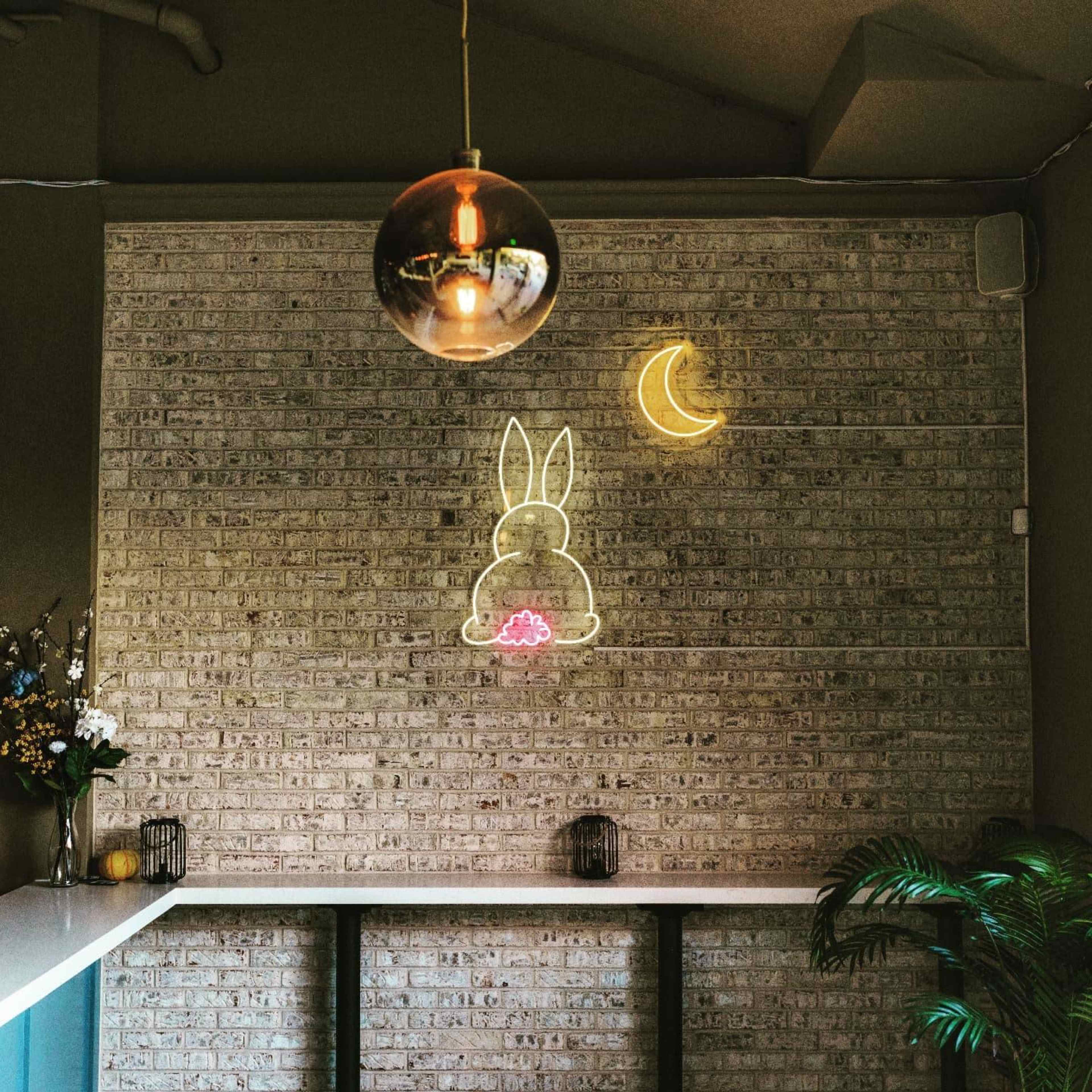 A brick wall features a neon bunny and crescent moon, illuminated by a hanging pendant light.