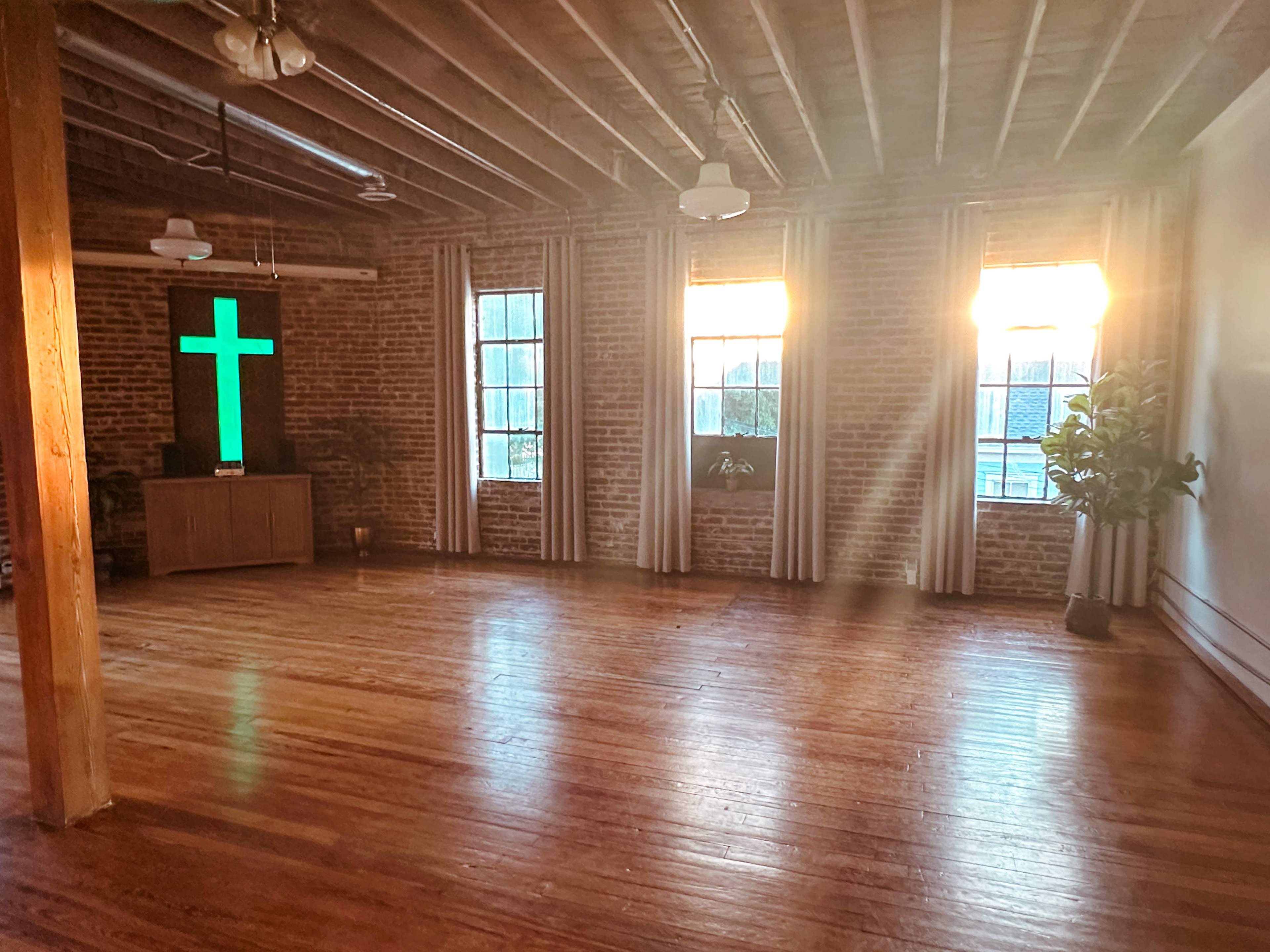 The image shows an empty room with wooden floors, exposed brick walls, large windows, and a green illuminated cross on a television screen.
