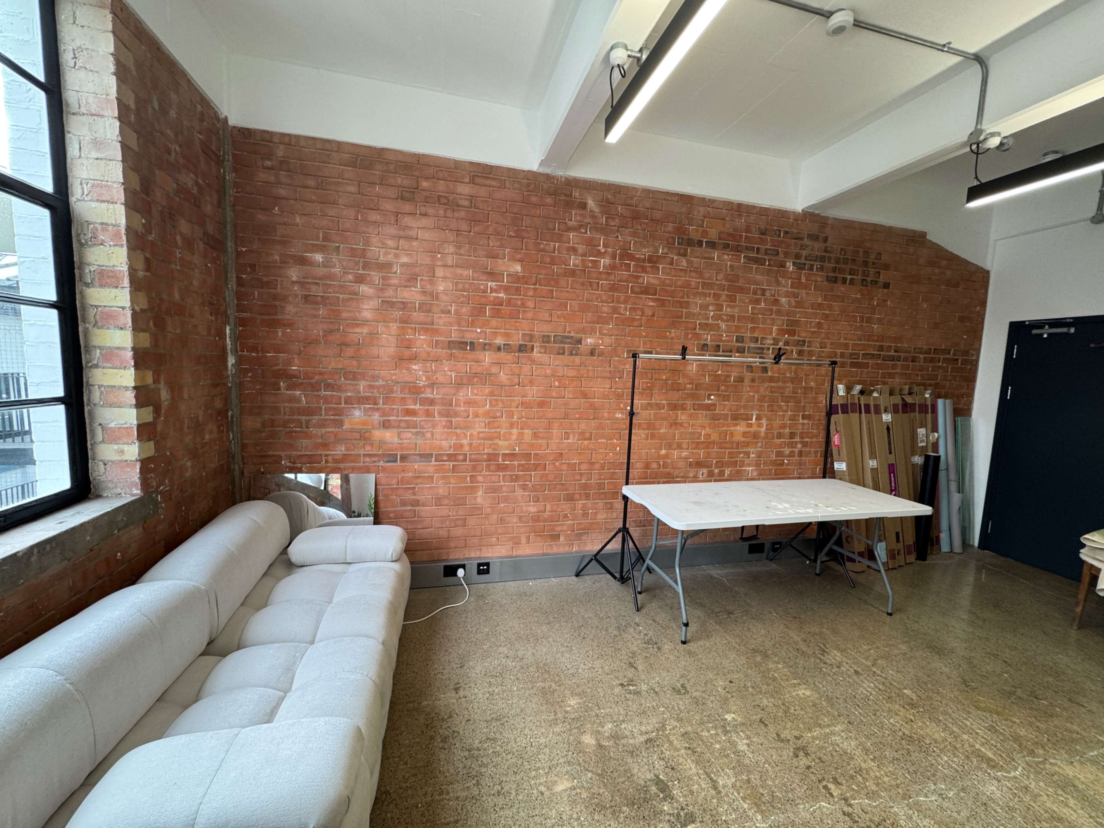 The image shows a spacious room with exposed brick walls, a light-colored sofa, a folding table, and wooden panels stacked against the wall.