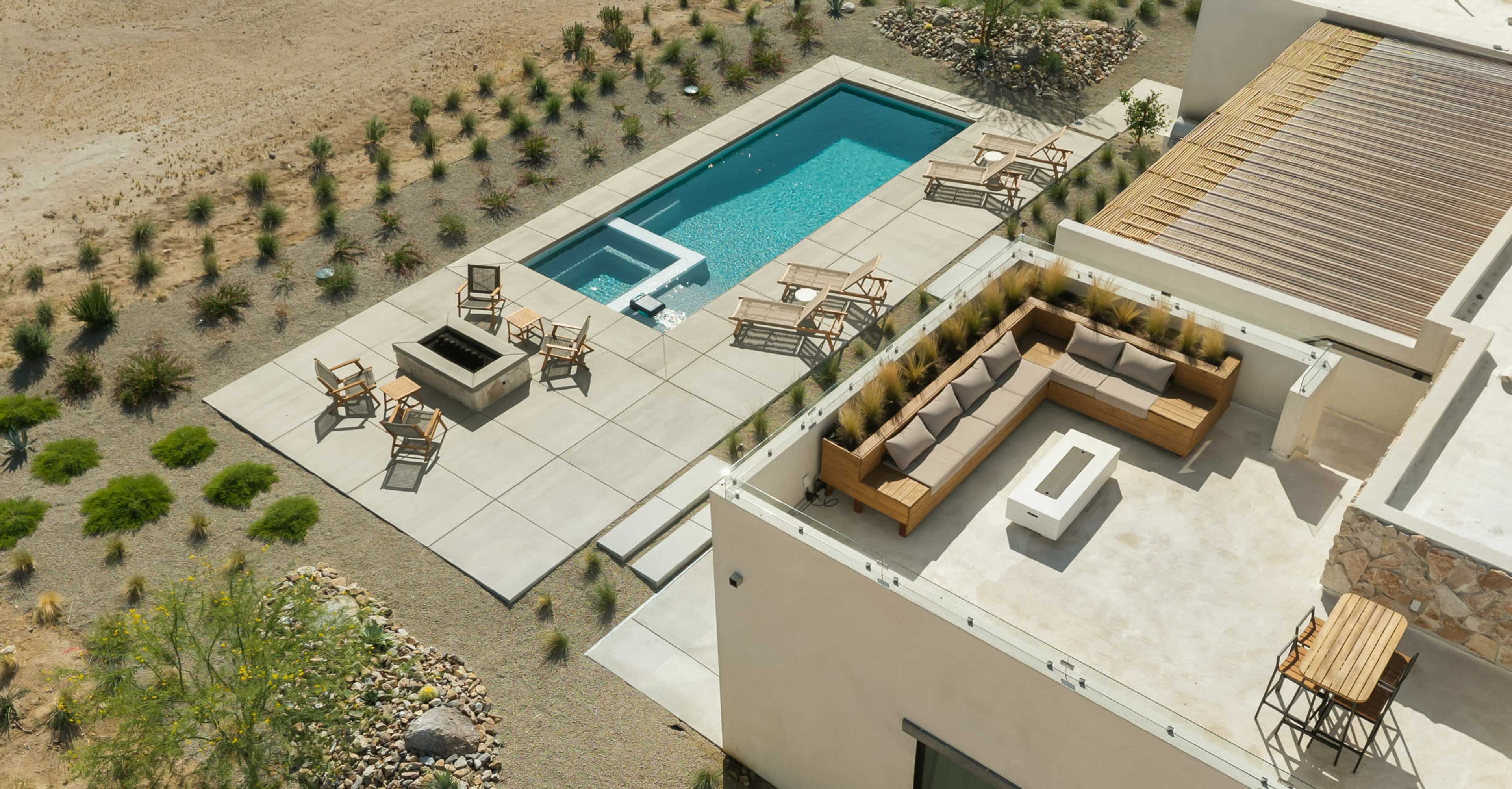 The image shows a modern patio area with a swimming pool, seating arrangements, and a fire pit, set in a desert landscape.