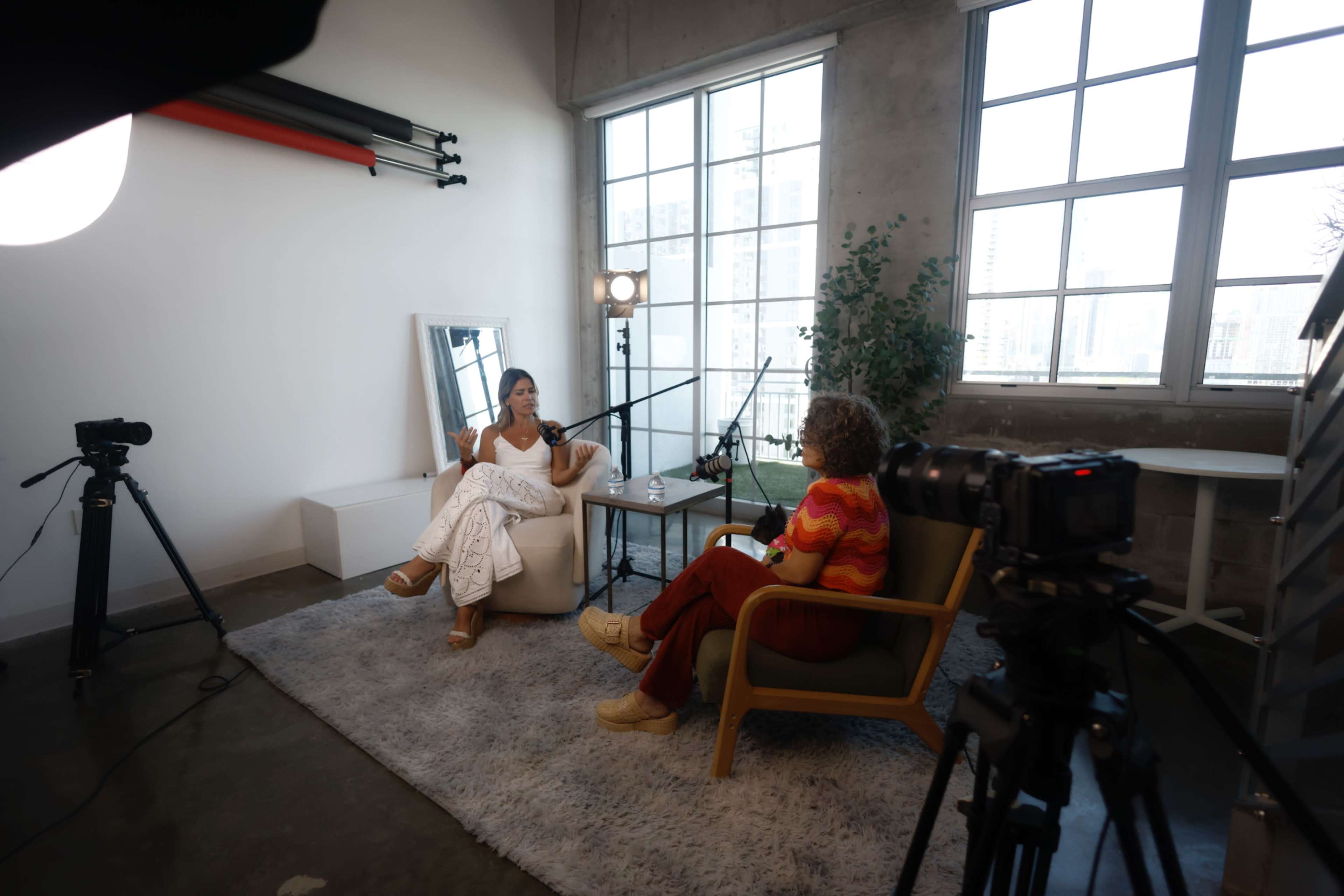 A woman in a white outfit sits in a chair while speaking to another woman in a colorful sweater, both positioned in a well-lit studio setting with large windows.