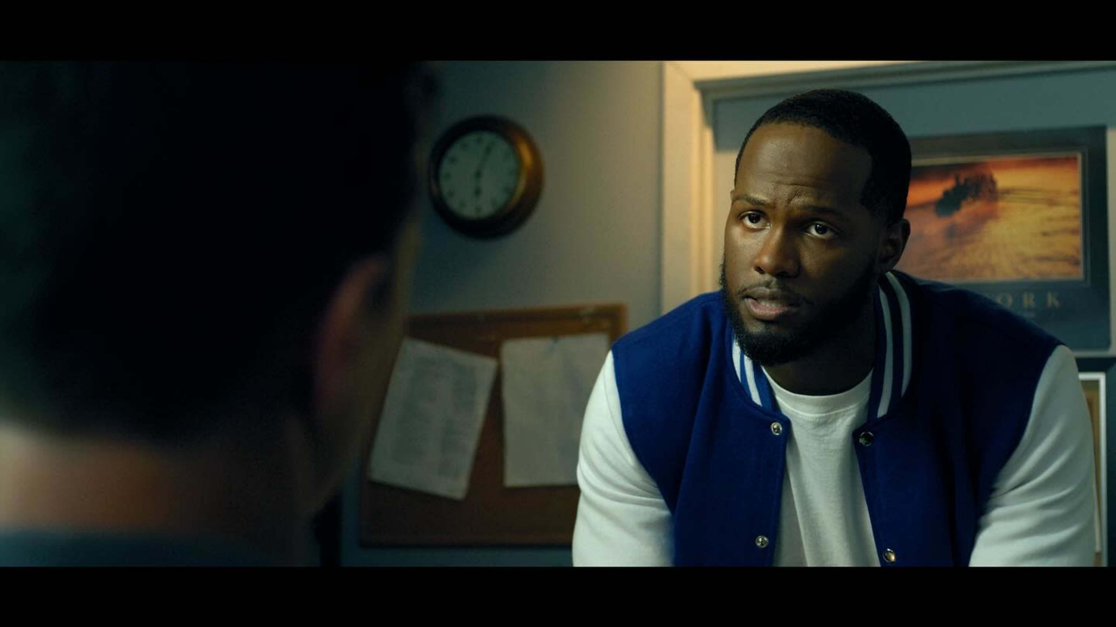 A man in a blue varsity jacket sits in an office, looking attentively at another person out of frame.