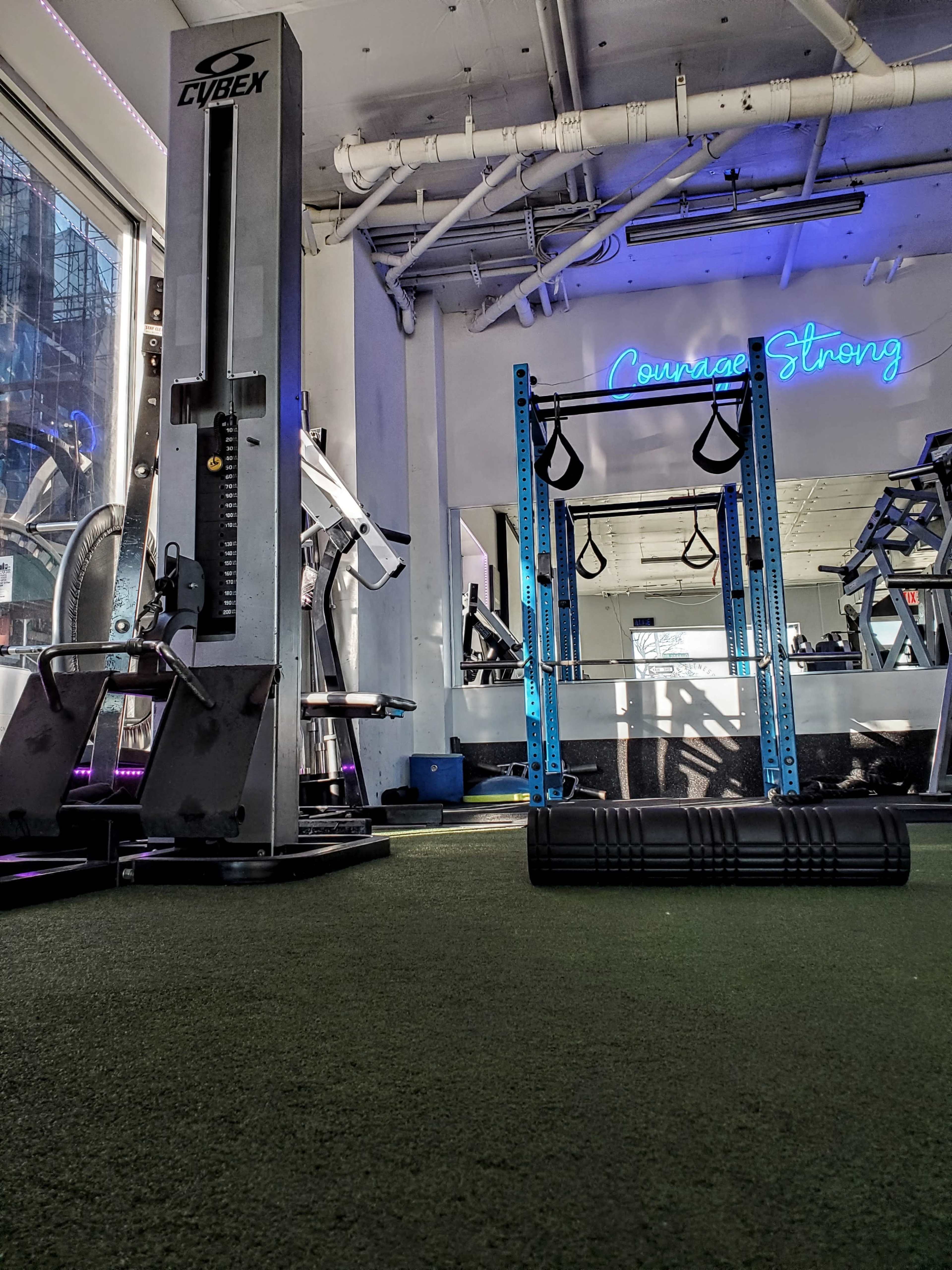 A gym interior with a Cybex machine on the left, a squat rack in the center, and exercise equipment in the background under a neon sign that says "Courage Strong."