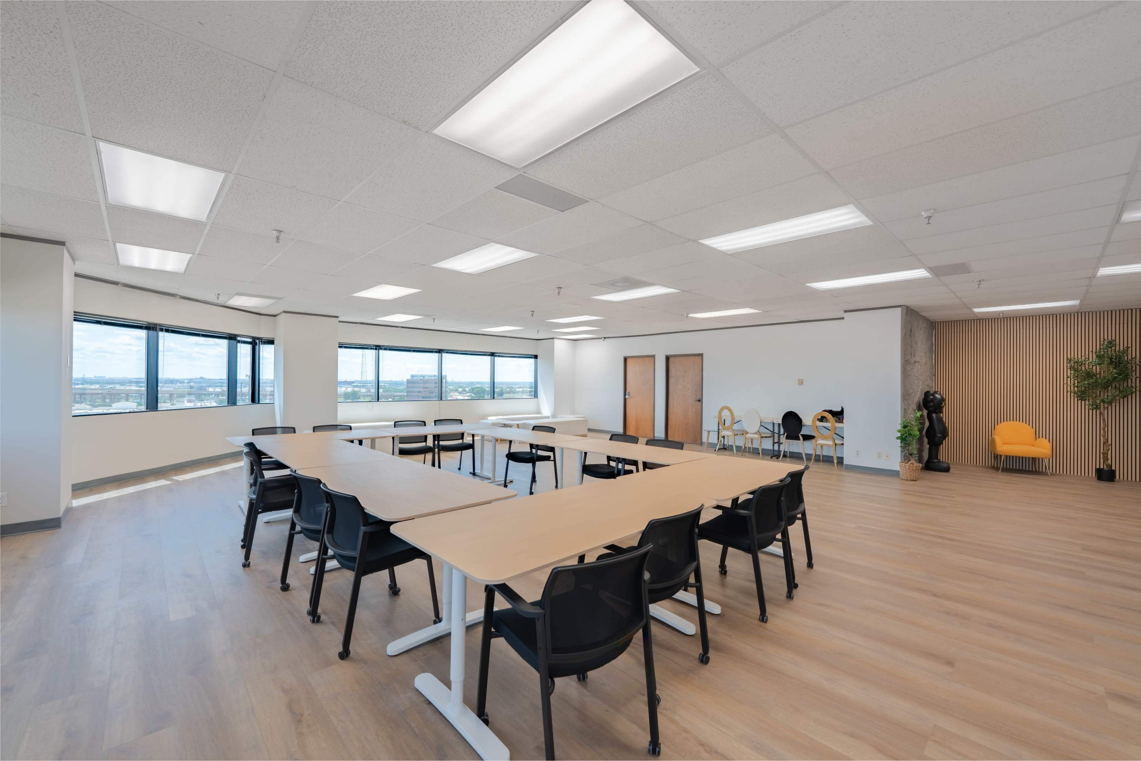 A large, bright meeting room features a long table surrounded by black chairs, with windows offering a view of the outside and minimalist decor on the walls.