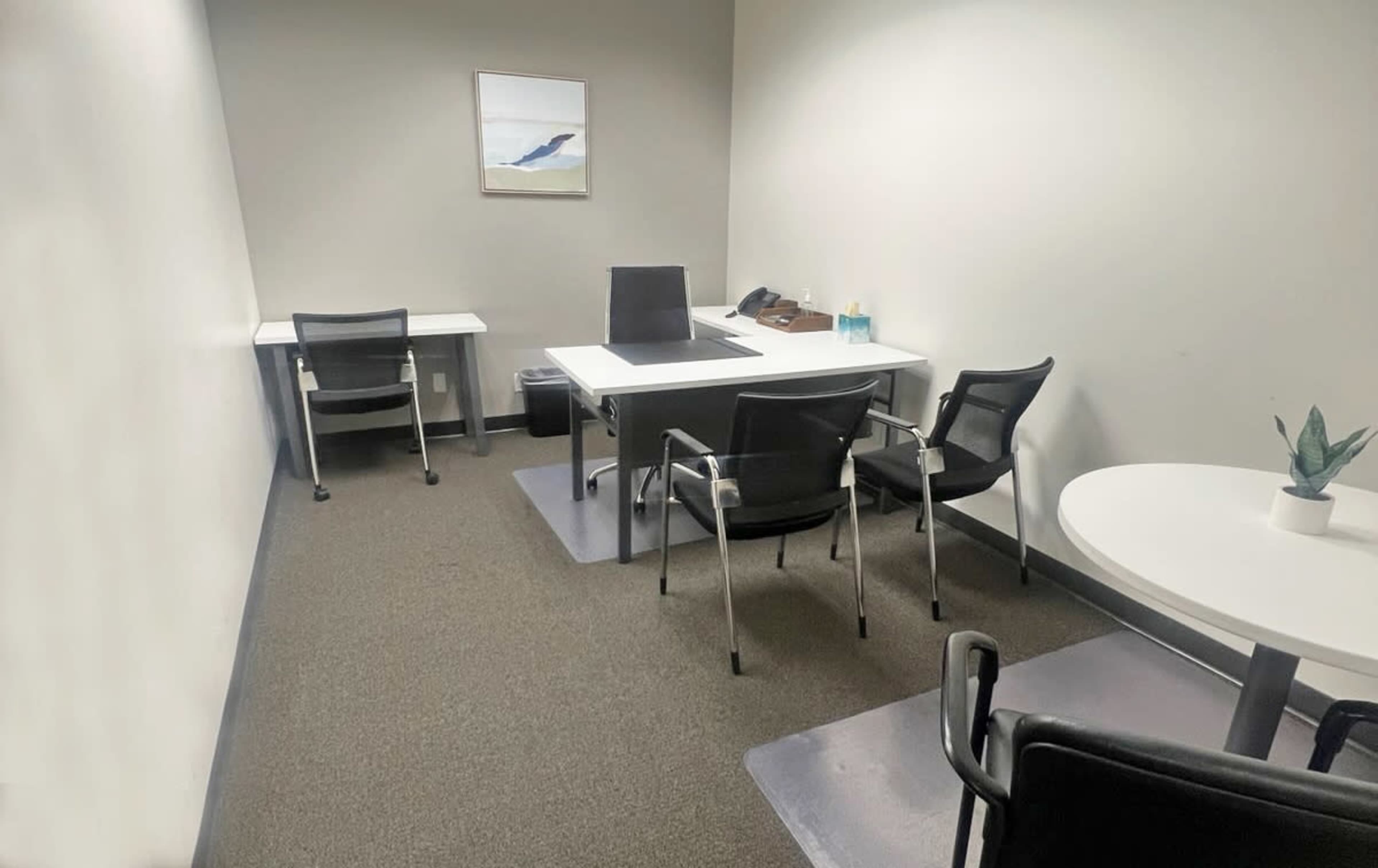 The image shows a small office space with two desks, several black chairs, and a round table, all set against light-colored walls.
