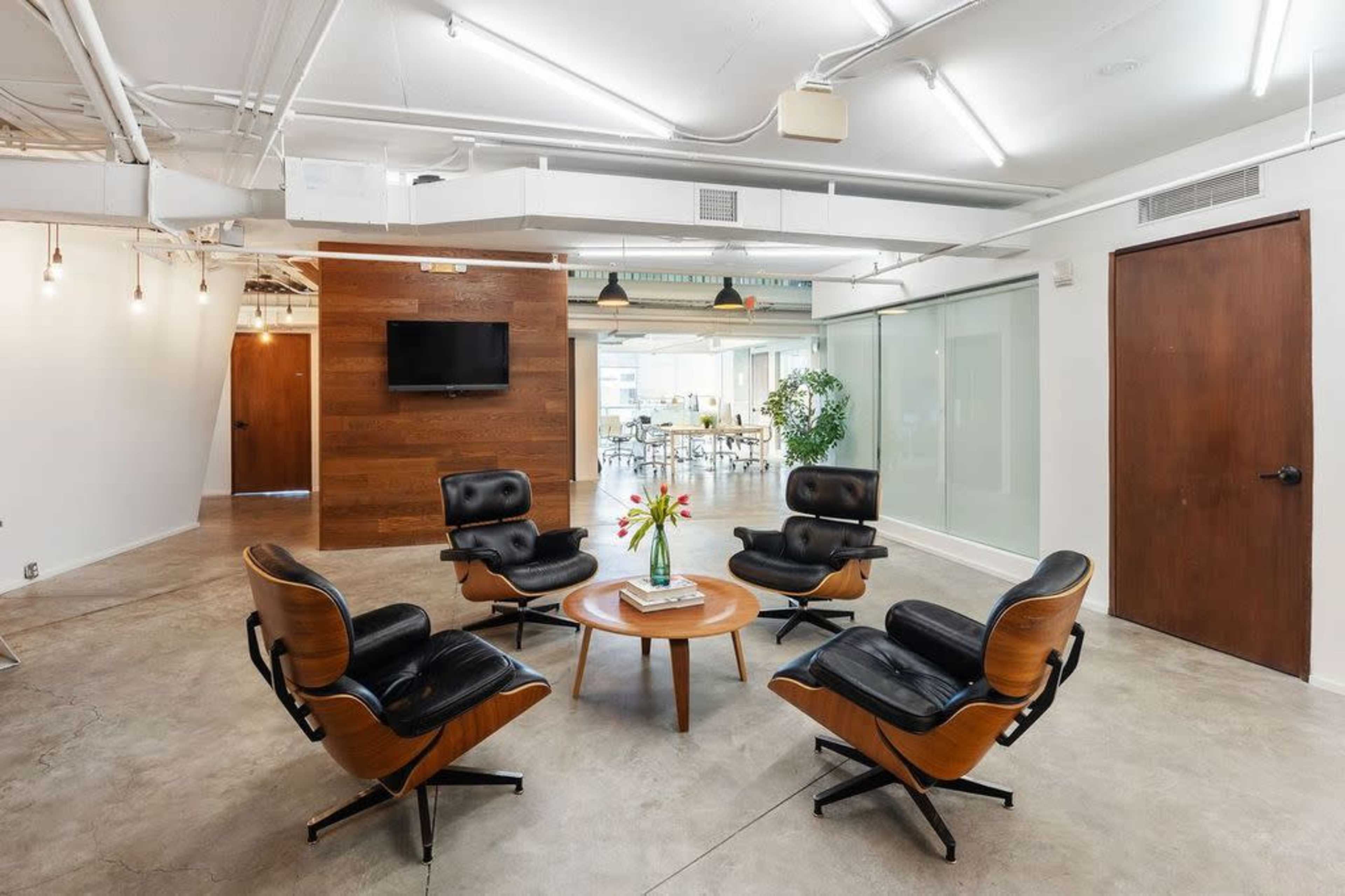 A modern office space features four black and wood lounge chairs arranged around a round table, with a potted plant and a glass wall in the background.