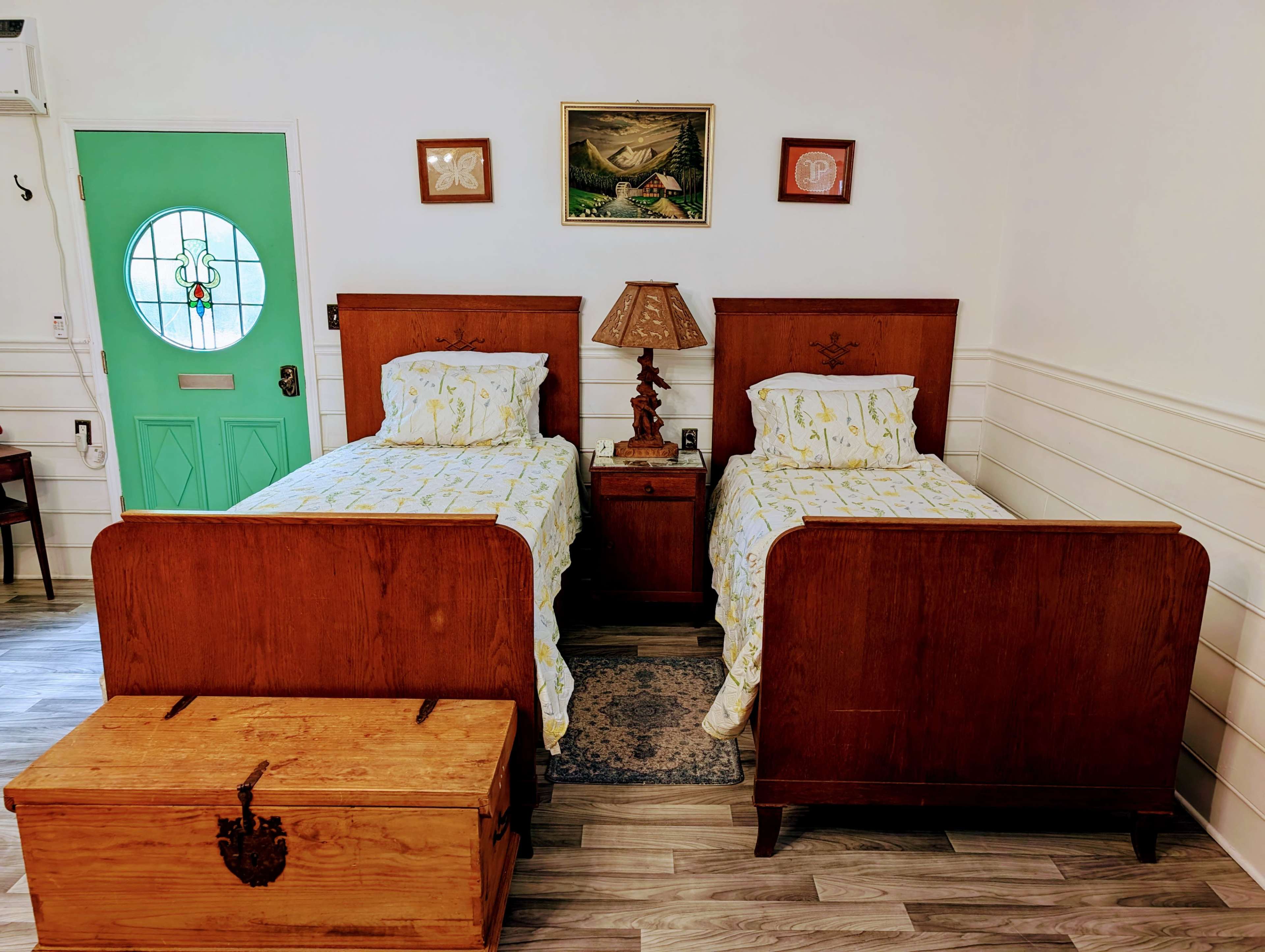 A room with two wooden twin beds with light yellow floral bedspread, a nightstand with a lamp between them, and a turquoise door in the background.