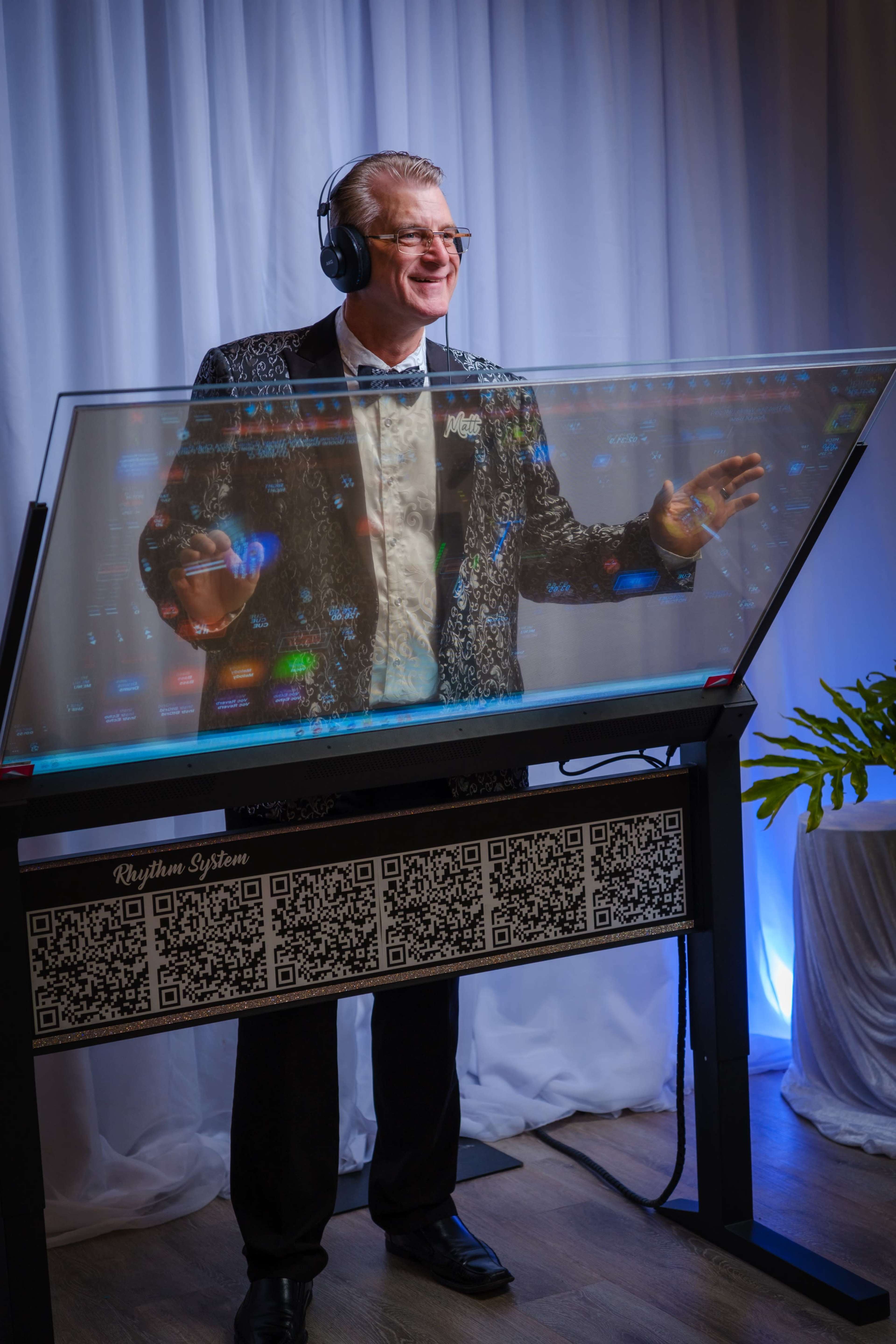 A man in a formal outfit, wearing headphones, stands behind a glass DJ booth with illuminated controls and a patterned background.