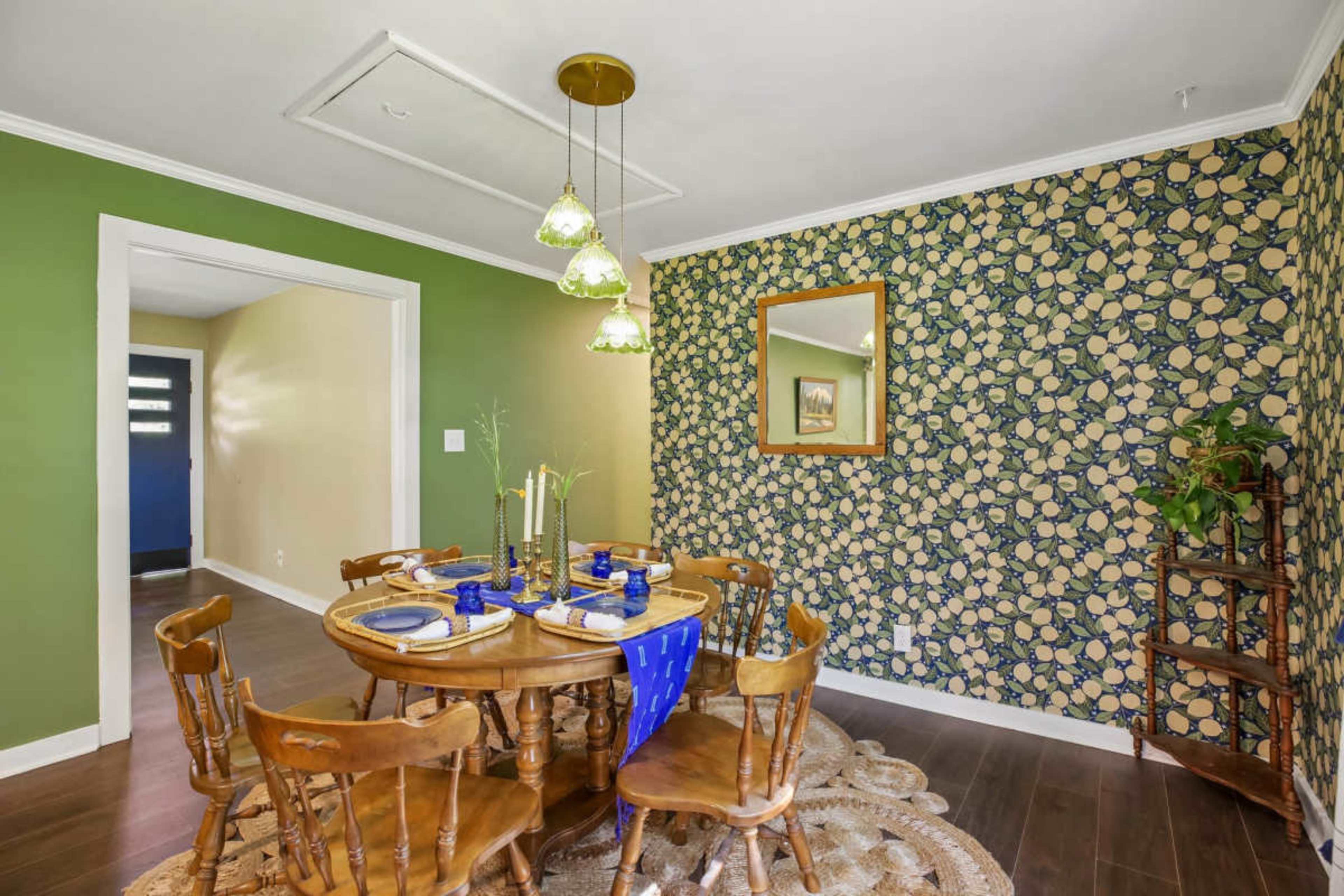 The image depicts a dining area with a round wooden table set for a meal, surrounded by eight chairs, against a floral wallpaper backdrop and green-painted walls.
