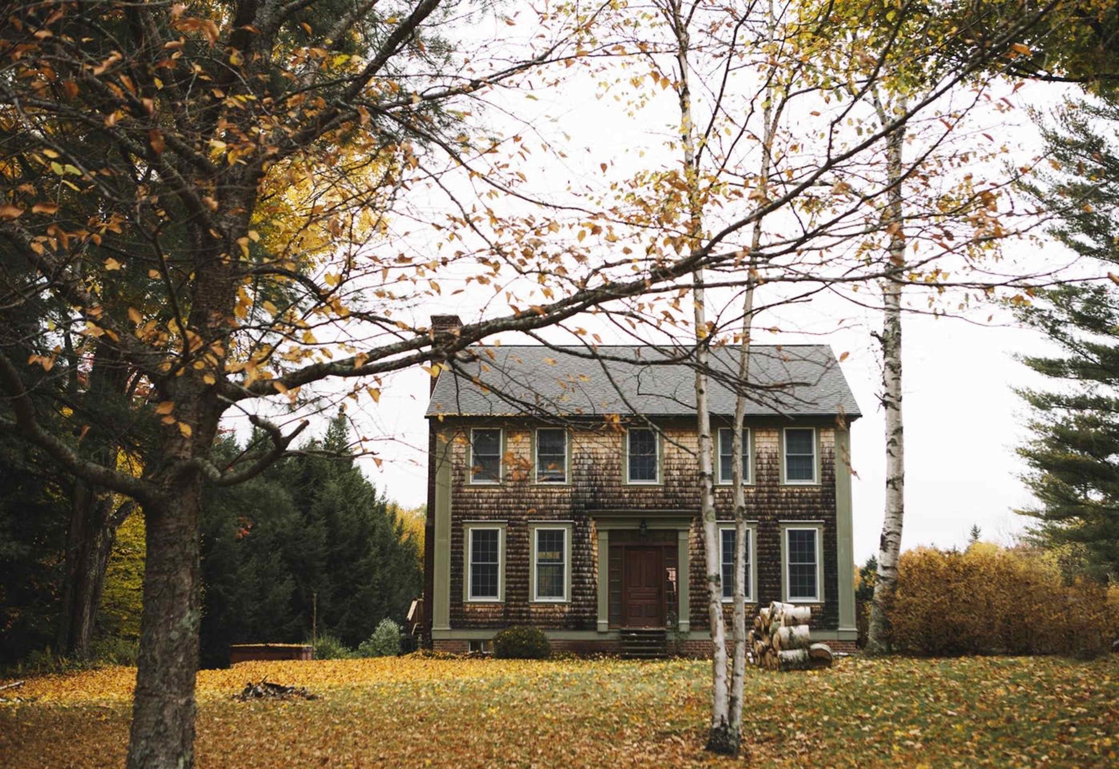 A two-story wooden house with a front porch is surrounded by trees showing fall foliage.