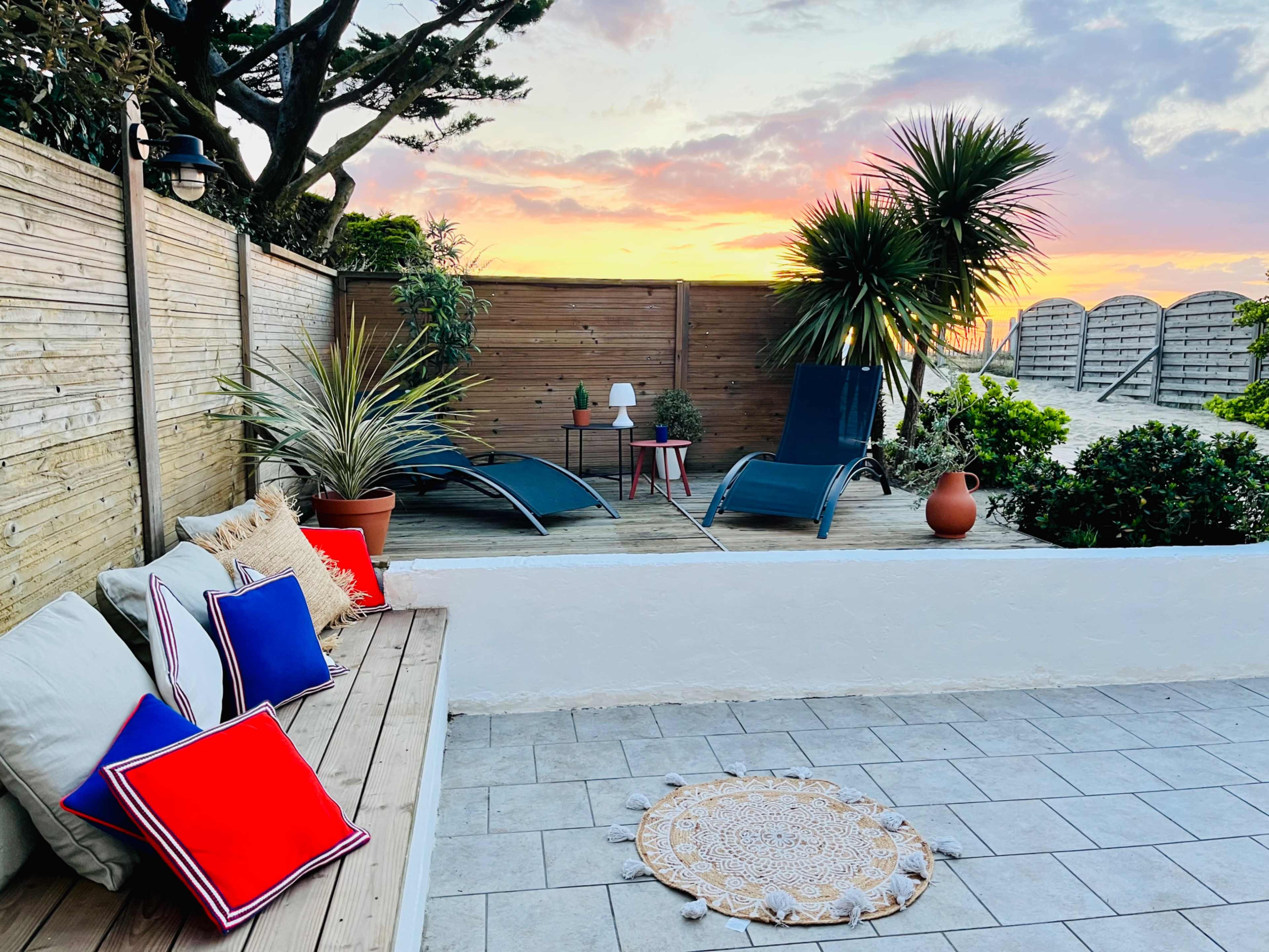 The image shows a cozy outdoor seating area with lounge chairs, potted plants, and decorative pillows, set against a colorful sunset sky.