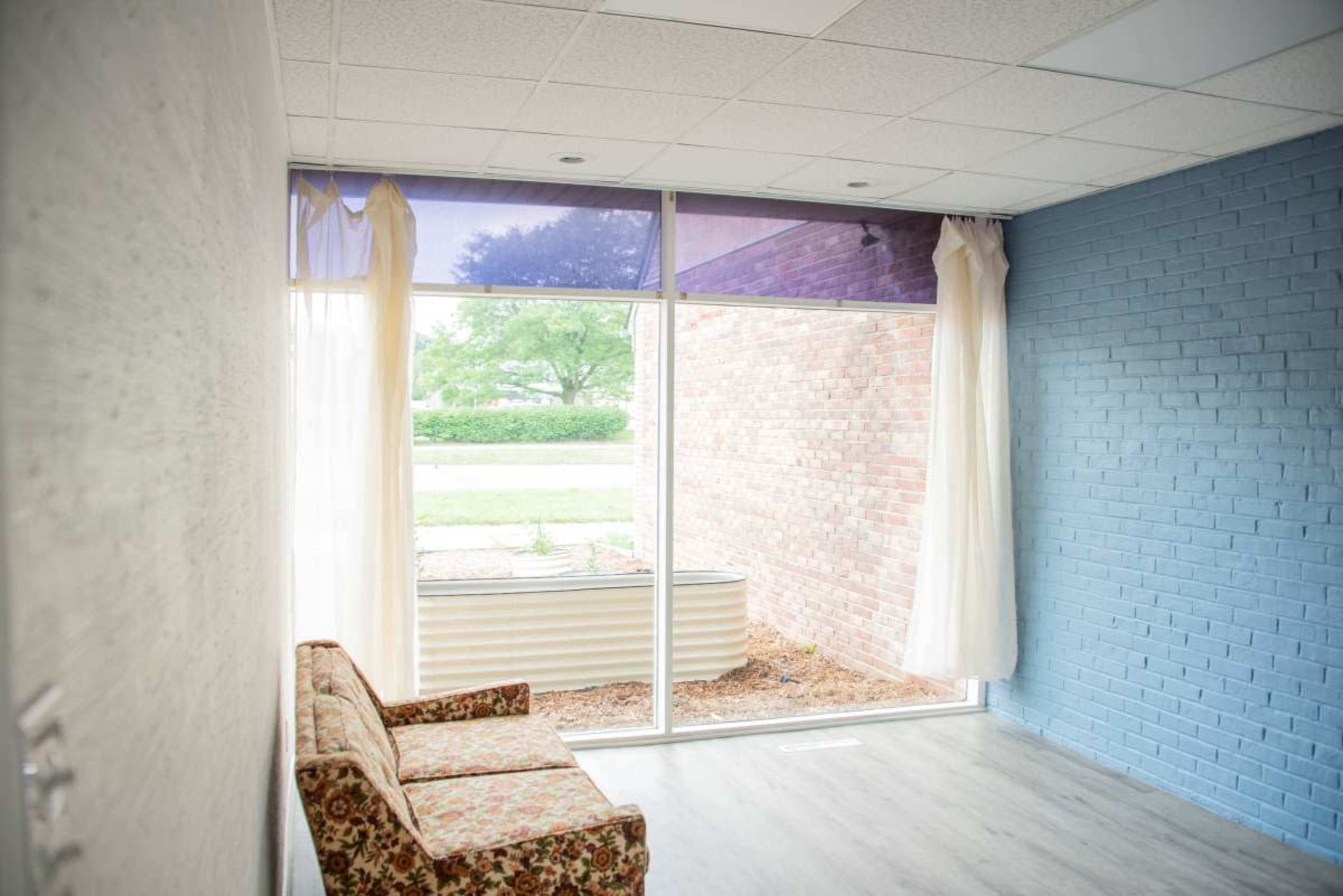 The image shows a small, empty room with a floral-patterned sofa and large windows overlooking a brick wall and outdoor greenery.