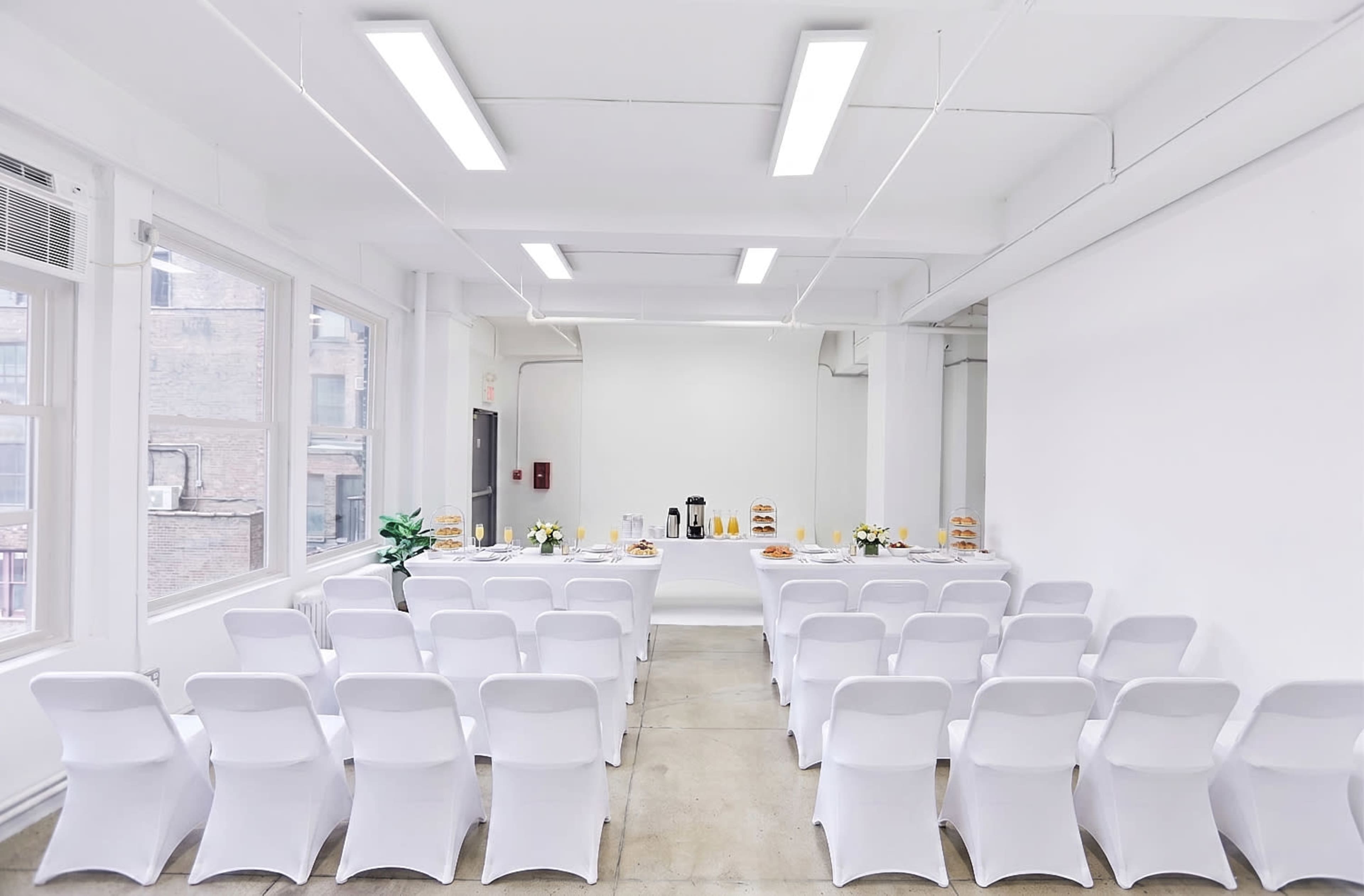 A bright, modern room features rows of white chairs facing a table set with refreshments, including coffee and pastries.