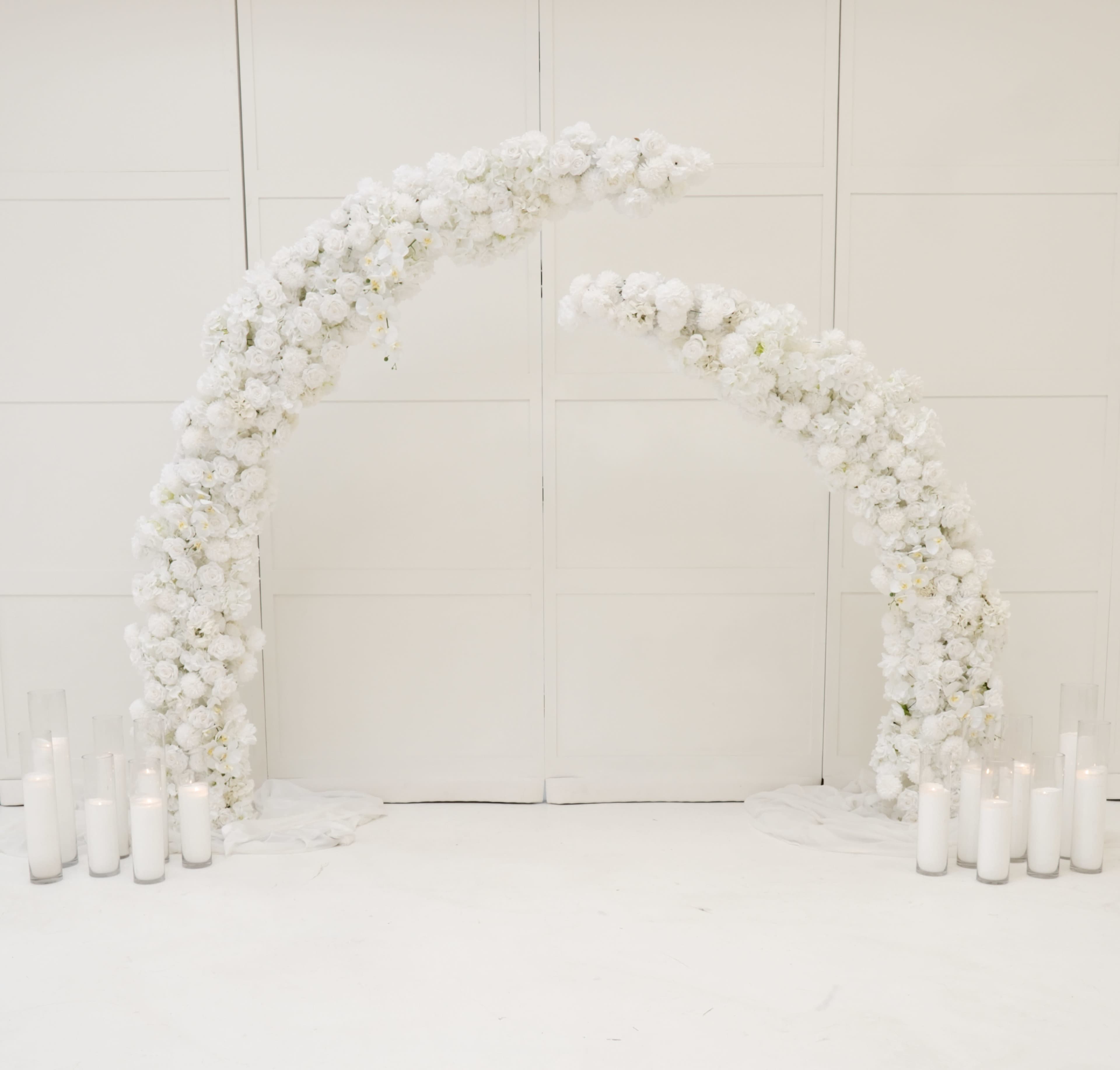The image shows a floral arch made of white flowers arranged in two curved structures, with white candles placed on the ground nearby.