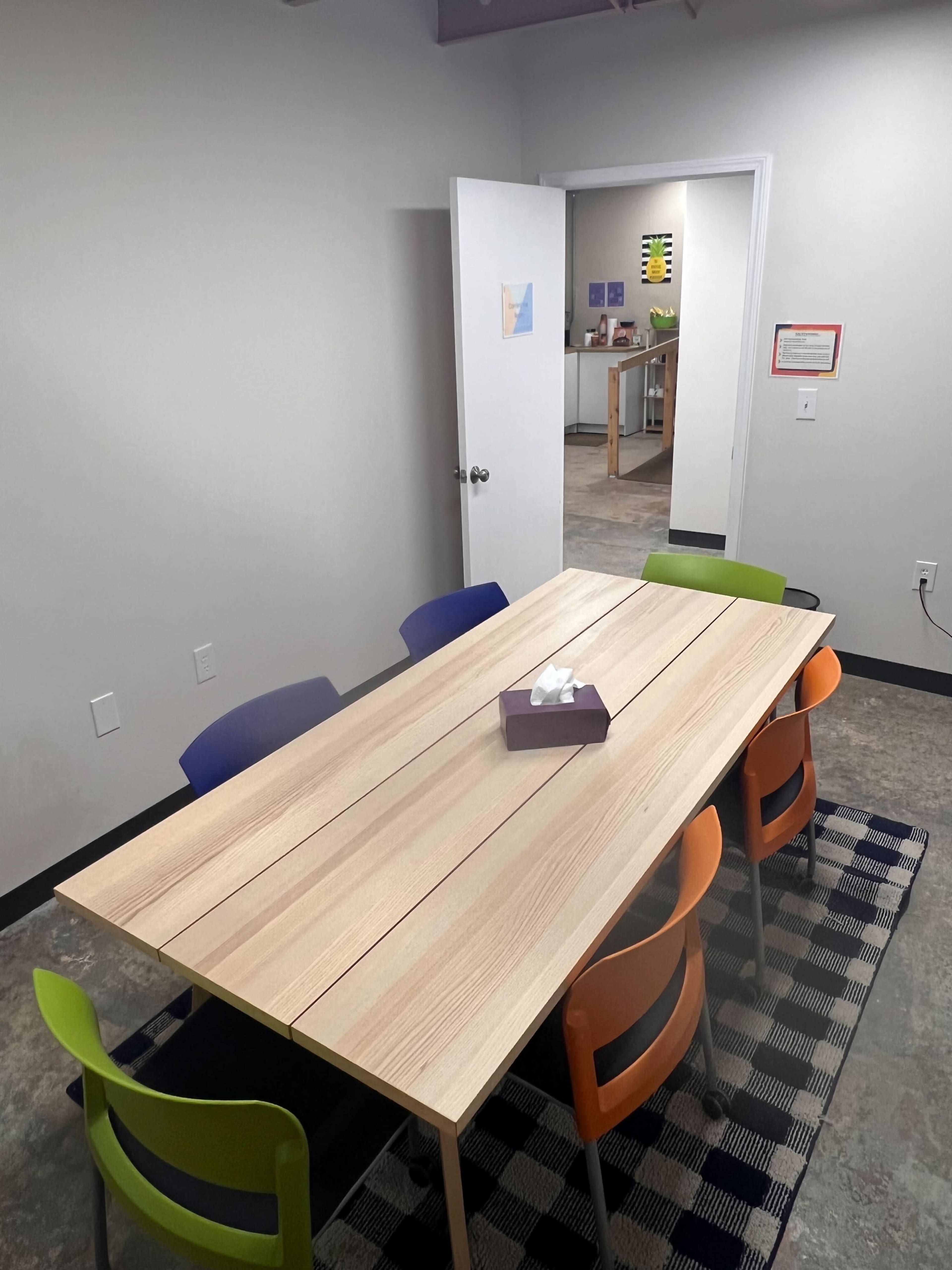A rectangular wooden table with a tissue box in the center is surrounded by colorful chairs in a meeting room, with a doorway leading to another area in the background.