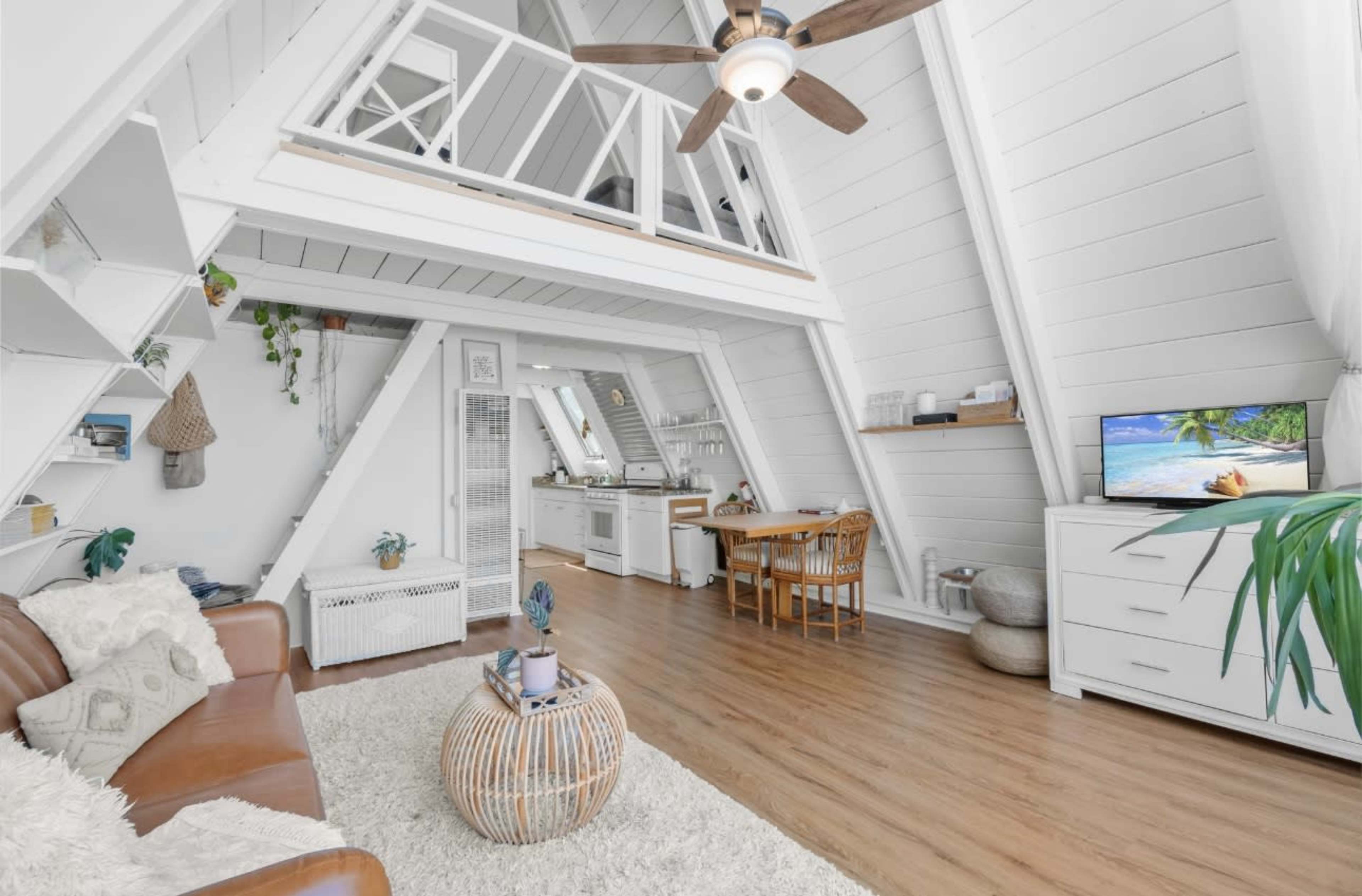 A bright, open-plan living space features a wooden floor, white walls, and an upstairs loft, along with a kitchen, dining area, and seating area.