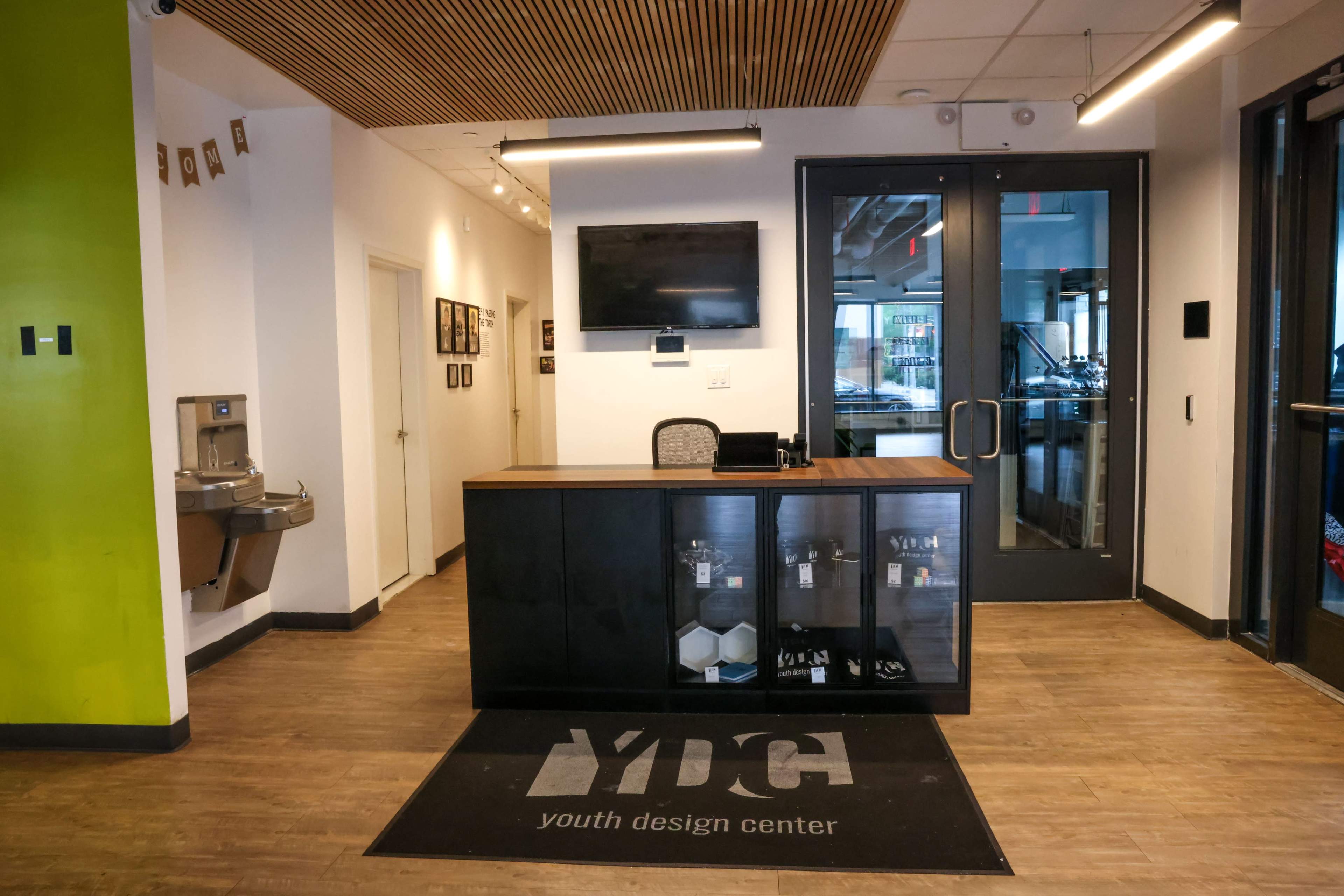 The image shows a reception area with a wooden front desk, a television mounted on the wall, a water fountain nearby, and double glass doors leading to another room.