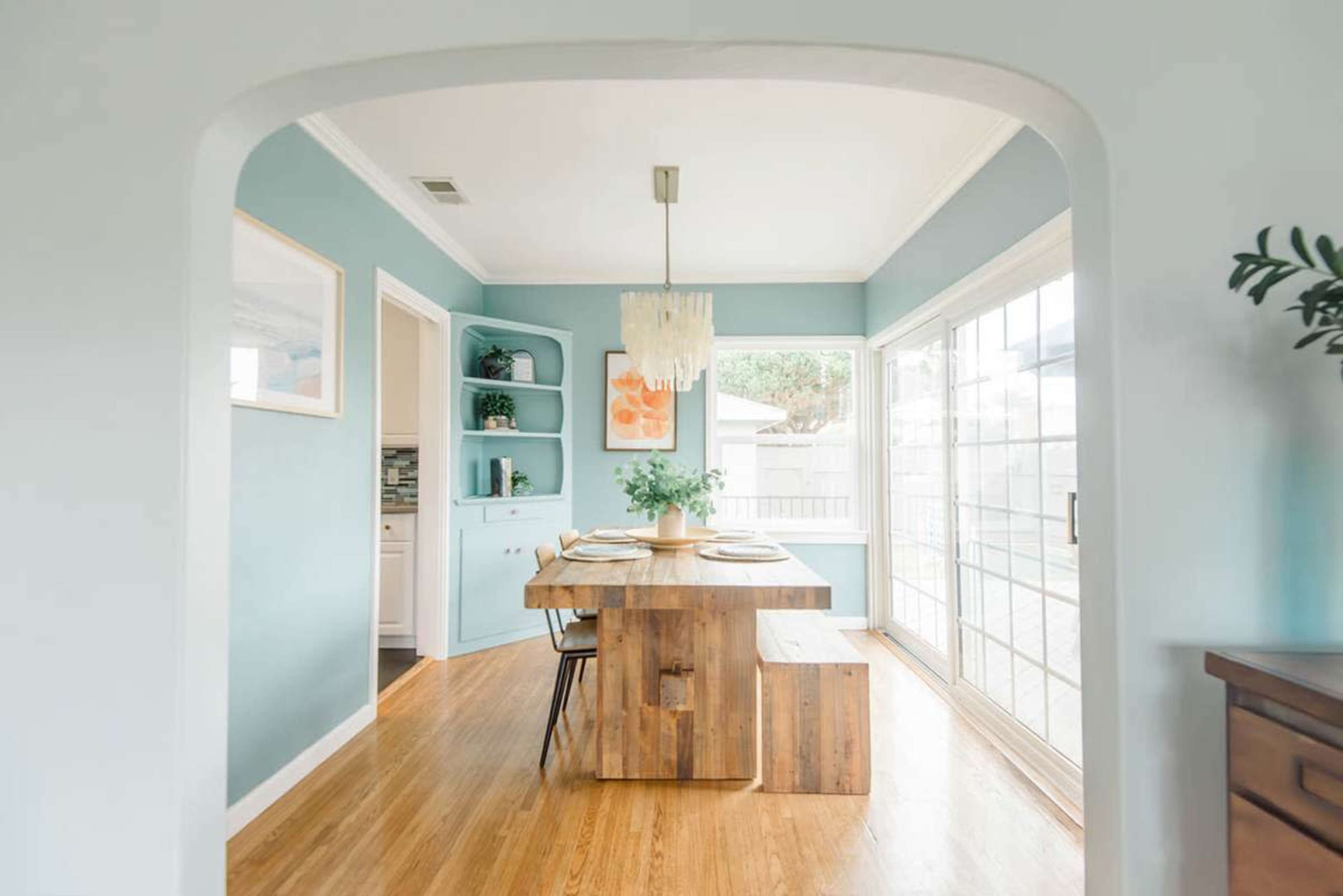 The image shows a dining area with a wooden table and chairs, surrounded by light blue walls and a large window that brings in natural light.