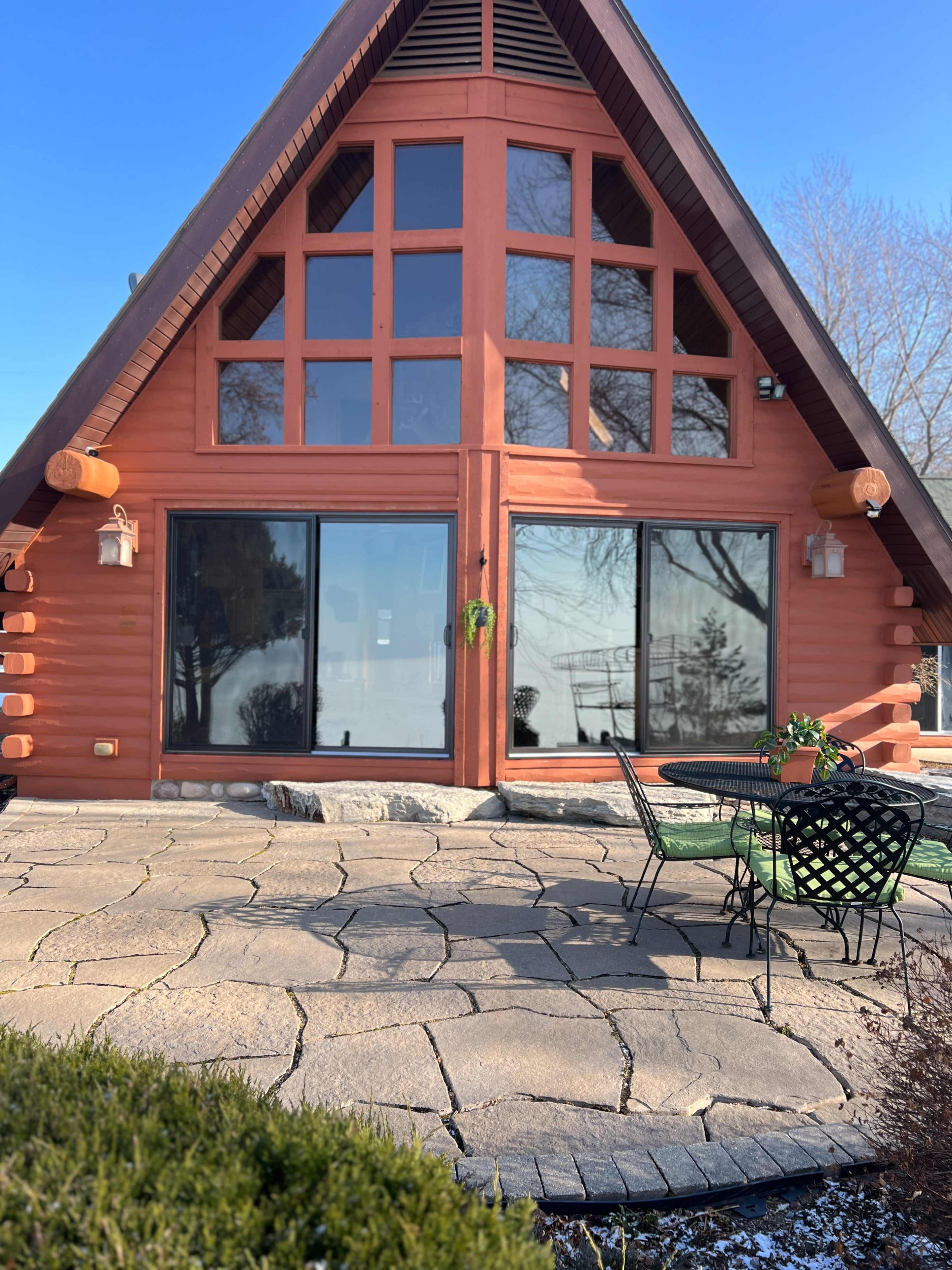 A triangular-shaped wooden cabin with large windows, surrounded by a stone patio and greenery.