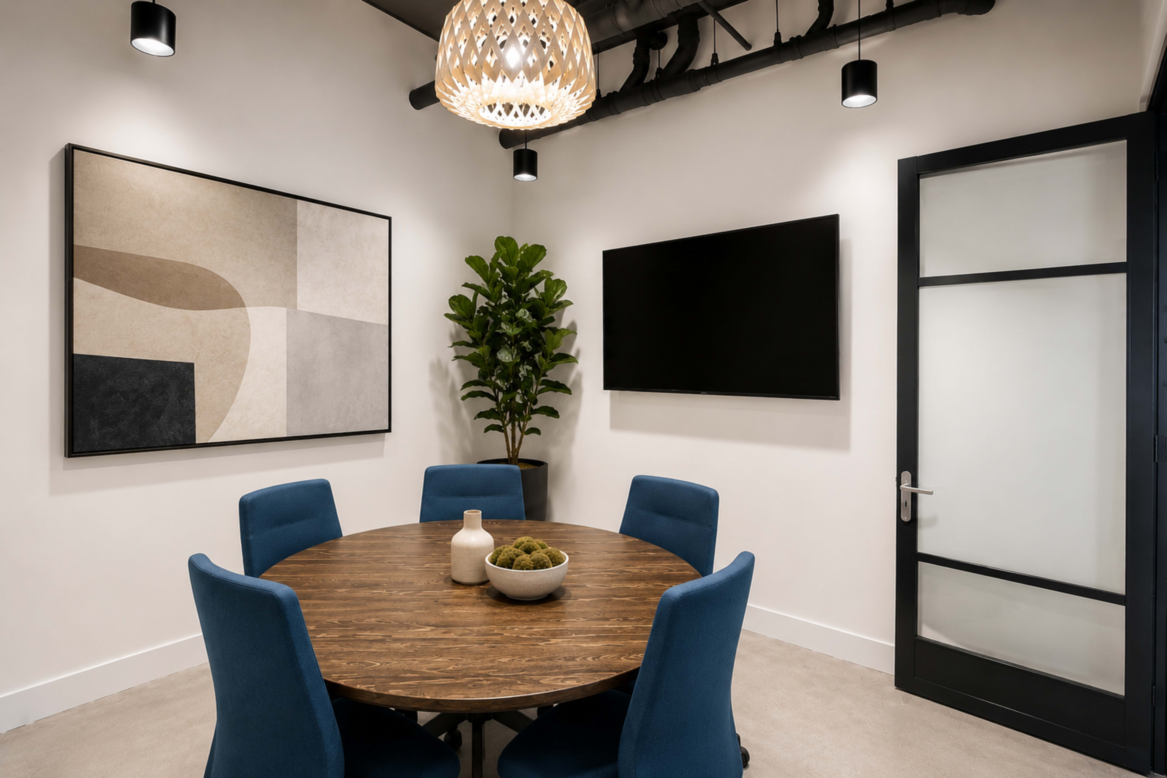 A modern meeting room features a round wooden table surrounded by blue chairs, a large abstract artwork on the wall, and a television mounted beside a glass door.