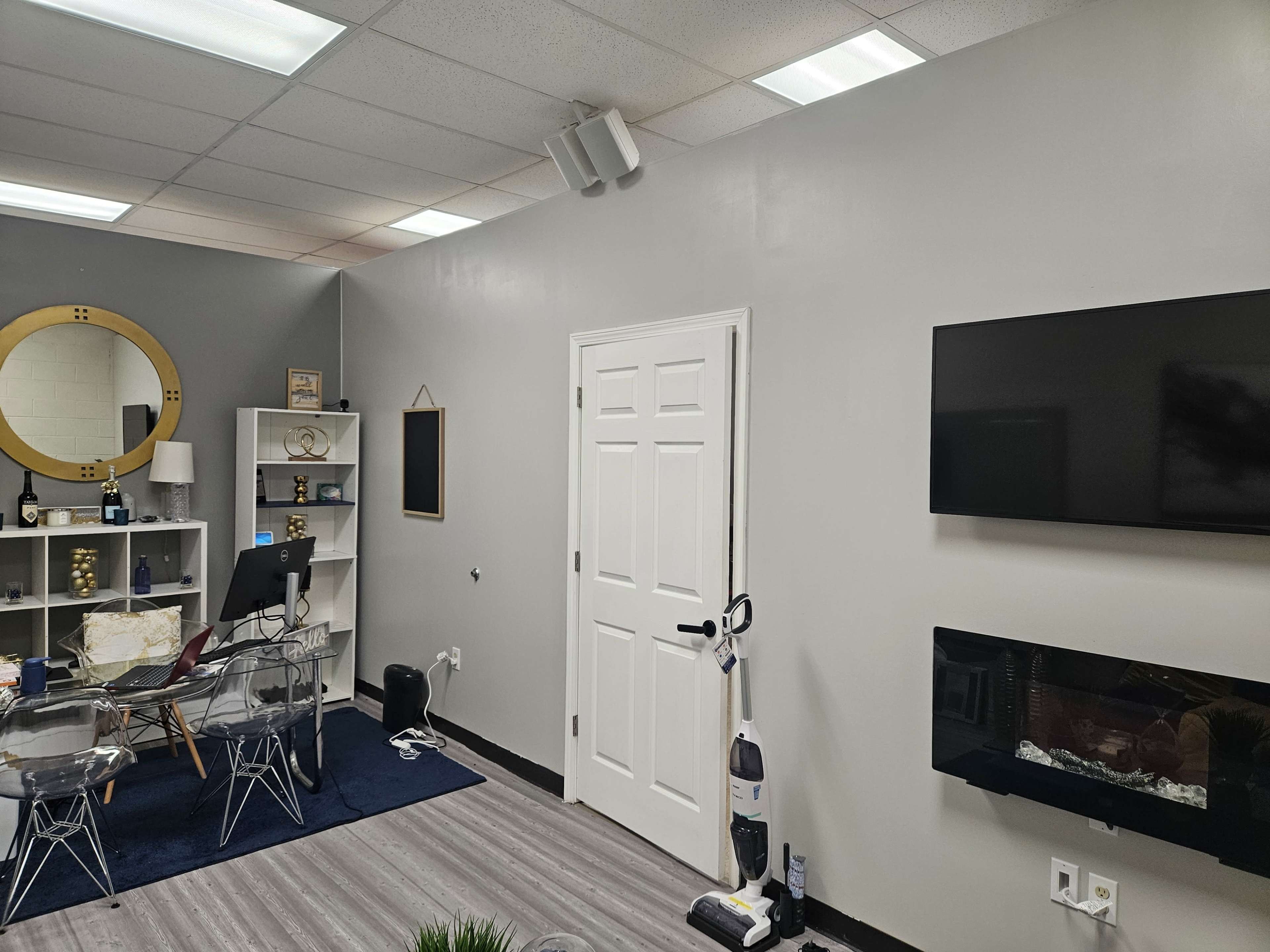 A modern office space features a TV mounted on a wall, a white door, a circular mirror, and transparent chairs arranged around a small table.