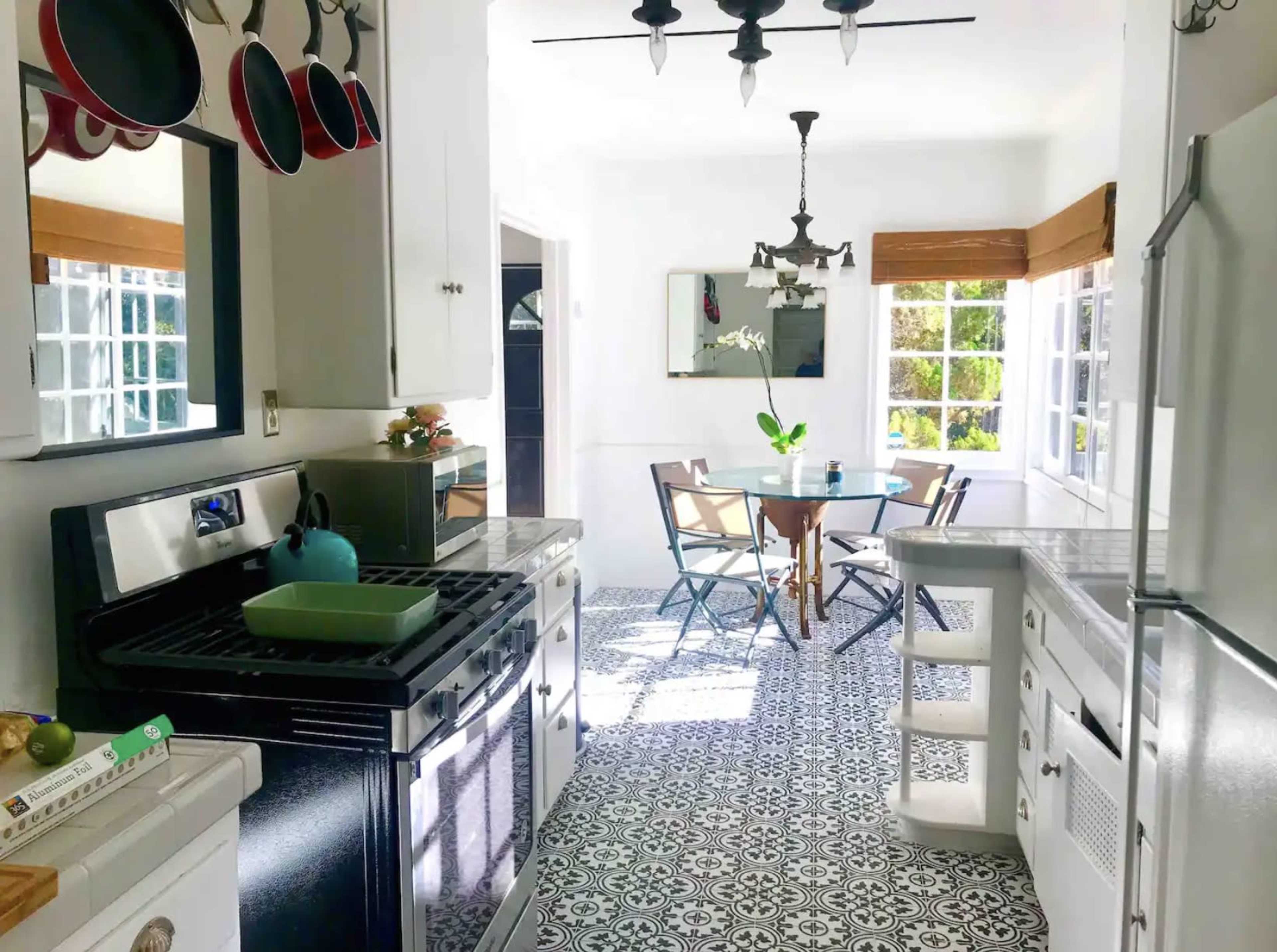 A bright kitchen features black appliances, white cabinets, and patterned floor tiles, with a dining area visible at the back.
