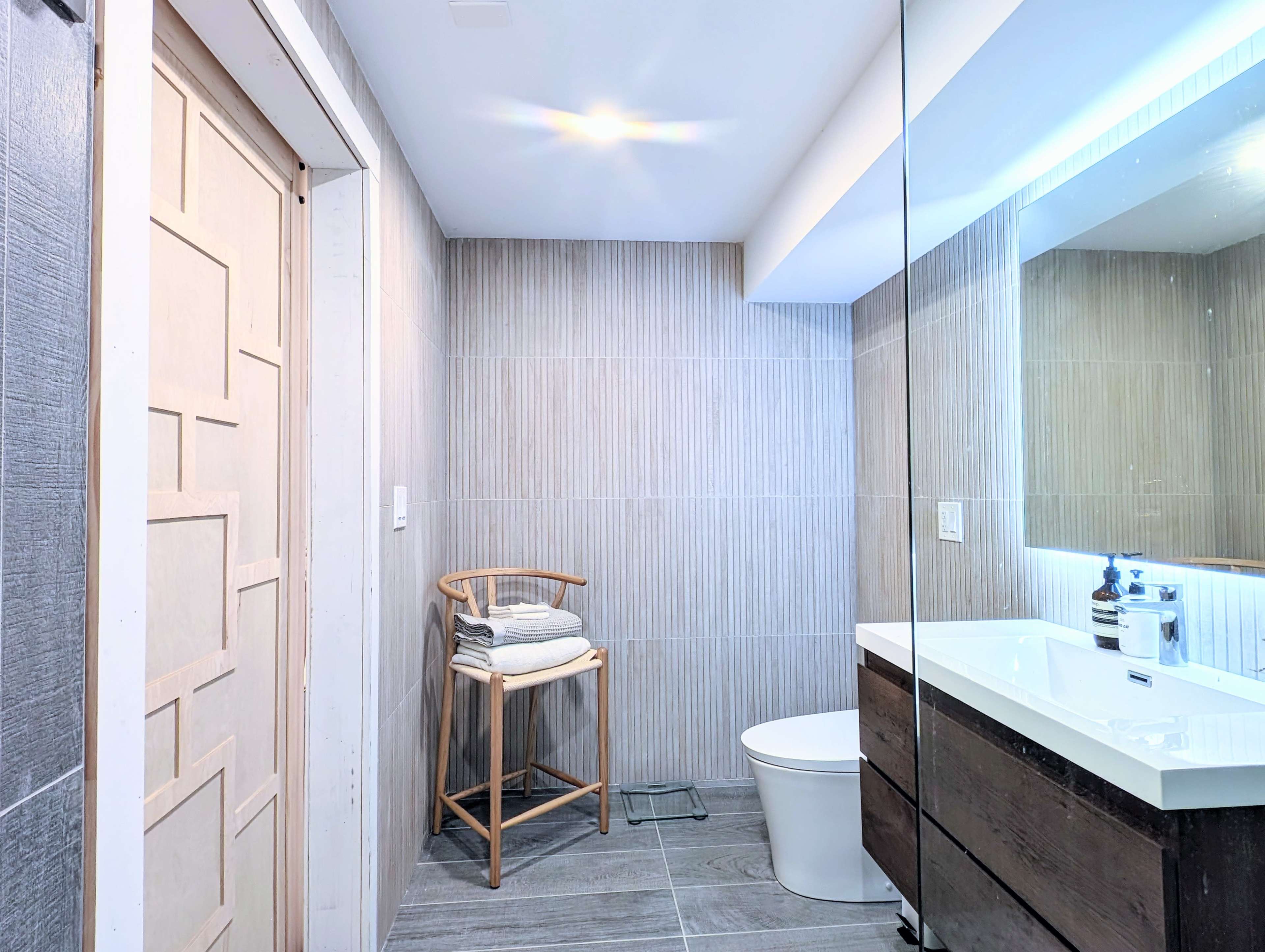 A modern bathroom with a wooden door, a wall-mounted mirror, a white sink, and a toilet, accompanied by a bamboo stool holding neatly stacked towels.