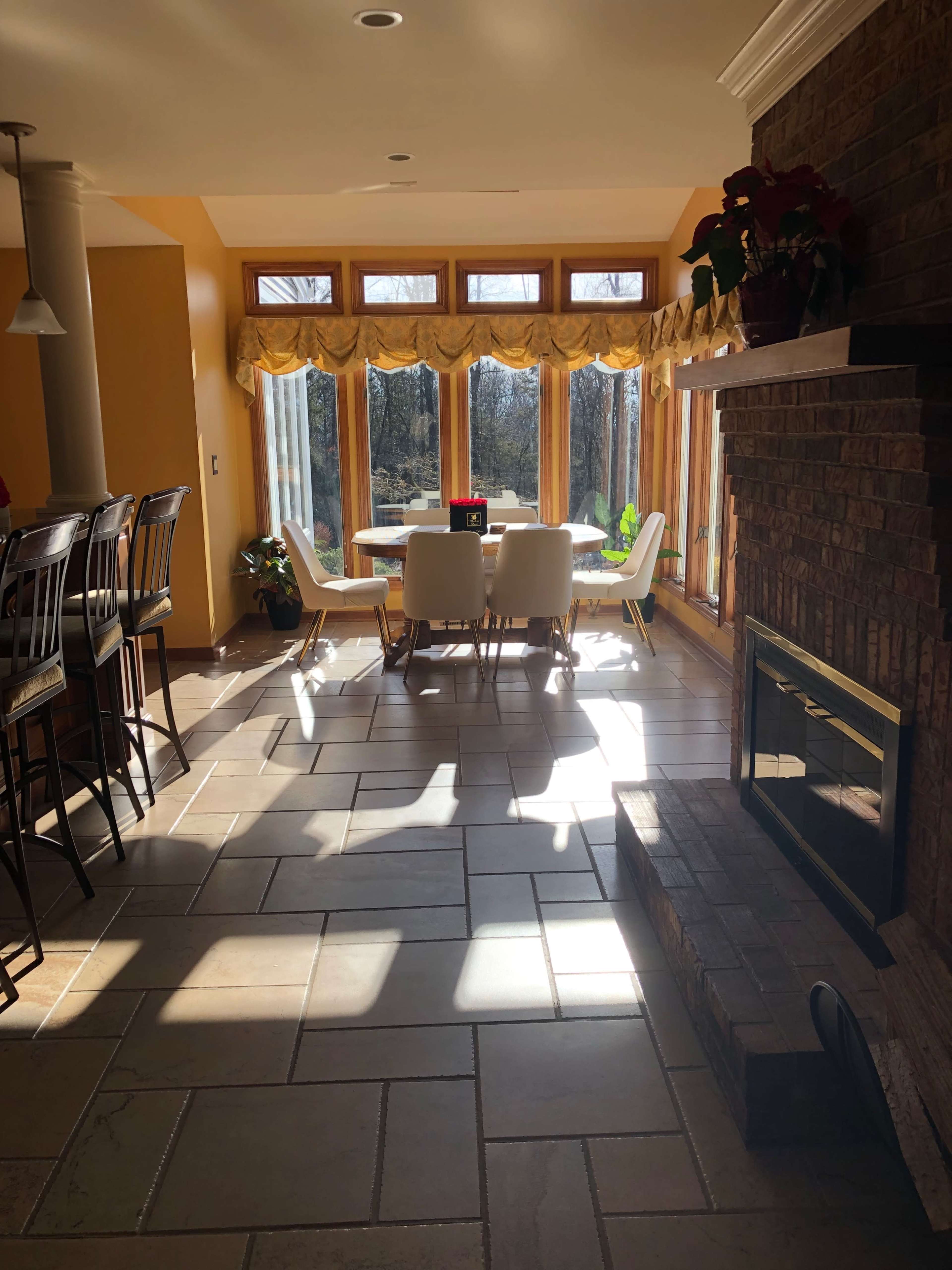 A sunlit dining area features a table surrounded by chairs, with large windows providing a view of the outdoors and a stone fireplace nearby.