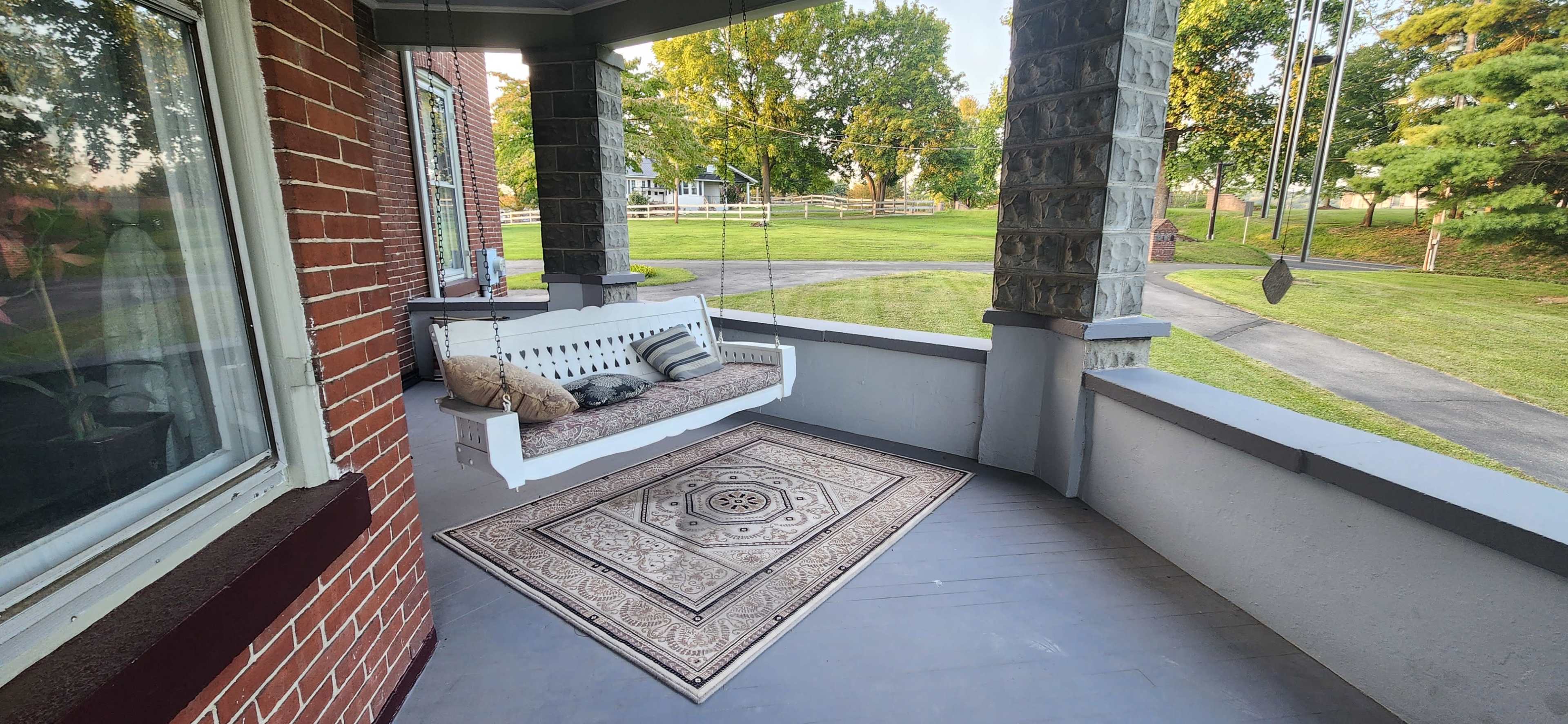 The image shows a porch with a hanging swing and a patterned rug, surrounded by greenery and a pathway.
