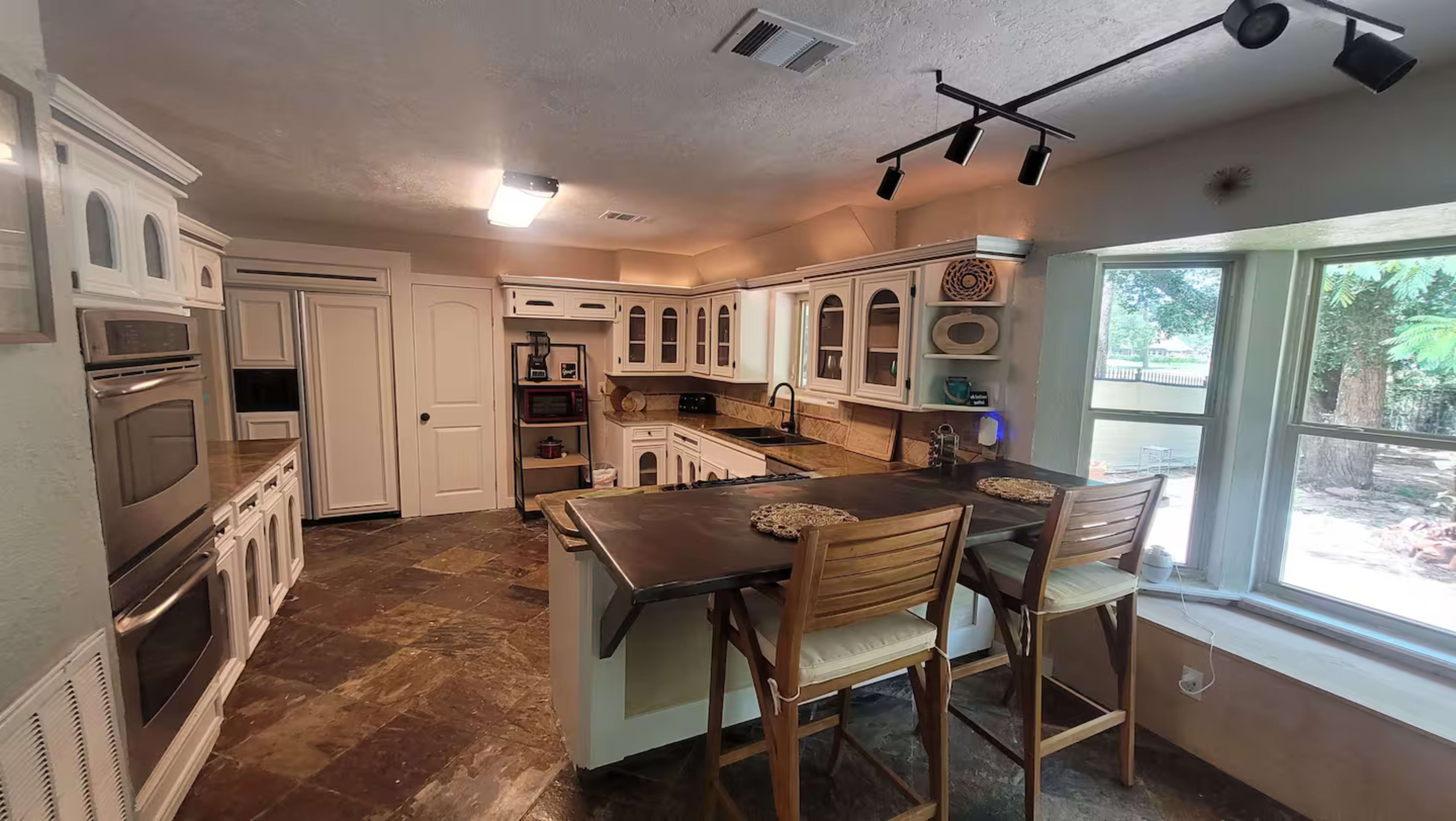 The image shows a well-lit kitchen featuring wooden cabinetry, a central island with bar stools, and a window view of the outdoors.