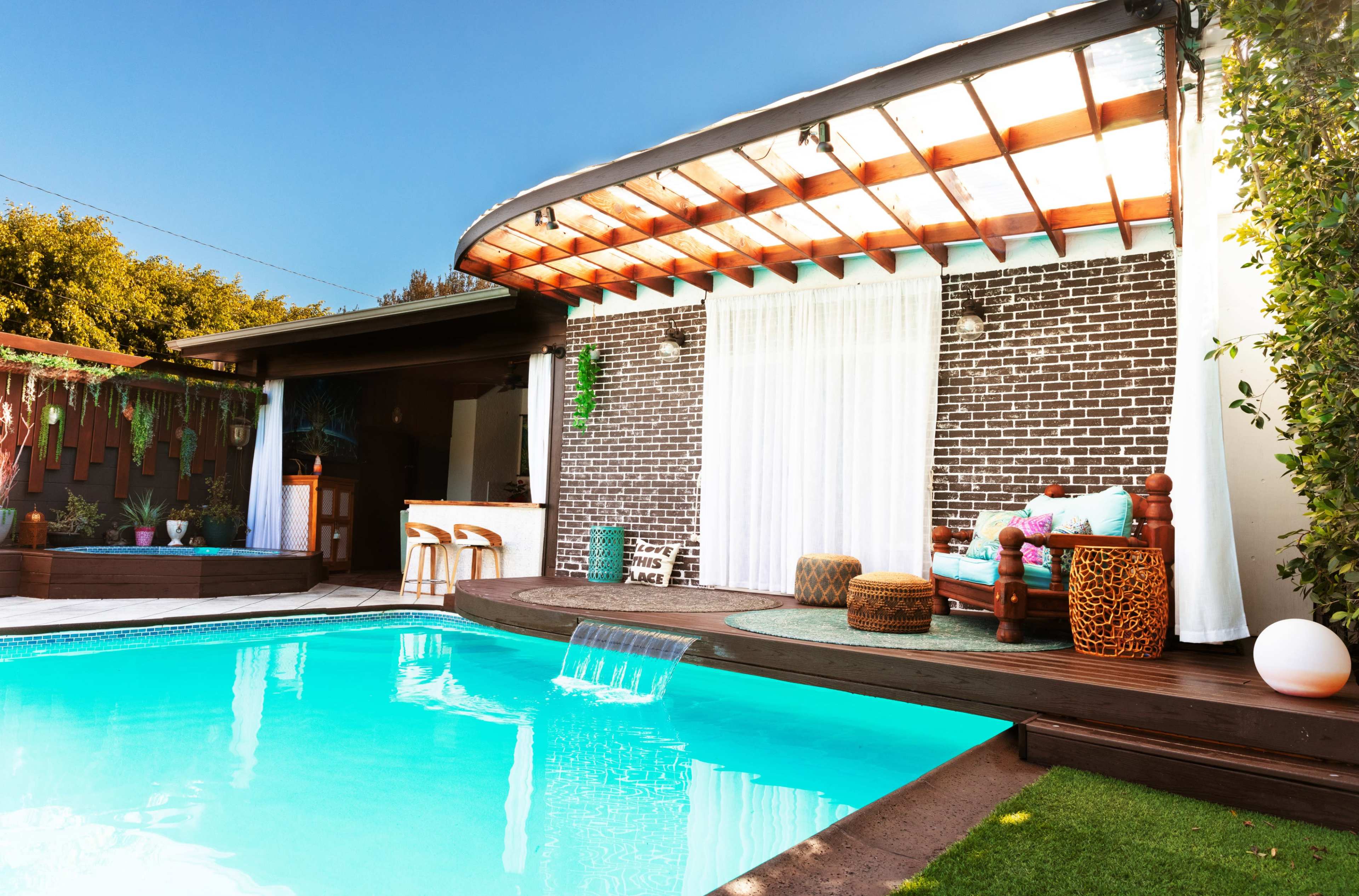 A modern backyard features a swimming pool with a water feature, surrounded by a patio and a brick wall, with comfortable seating and greenery.