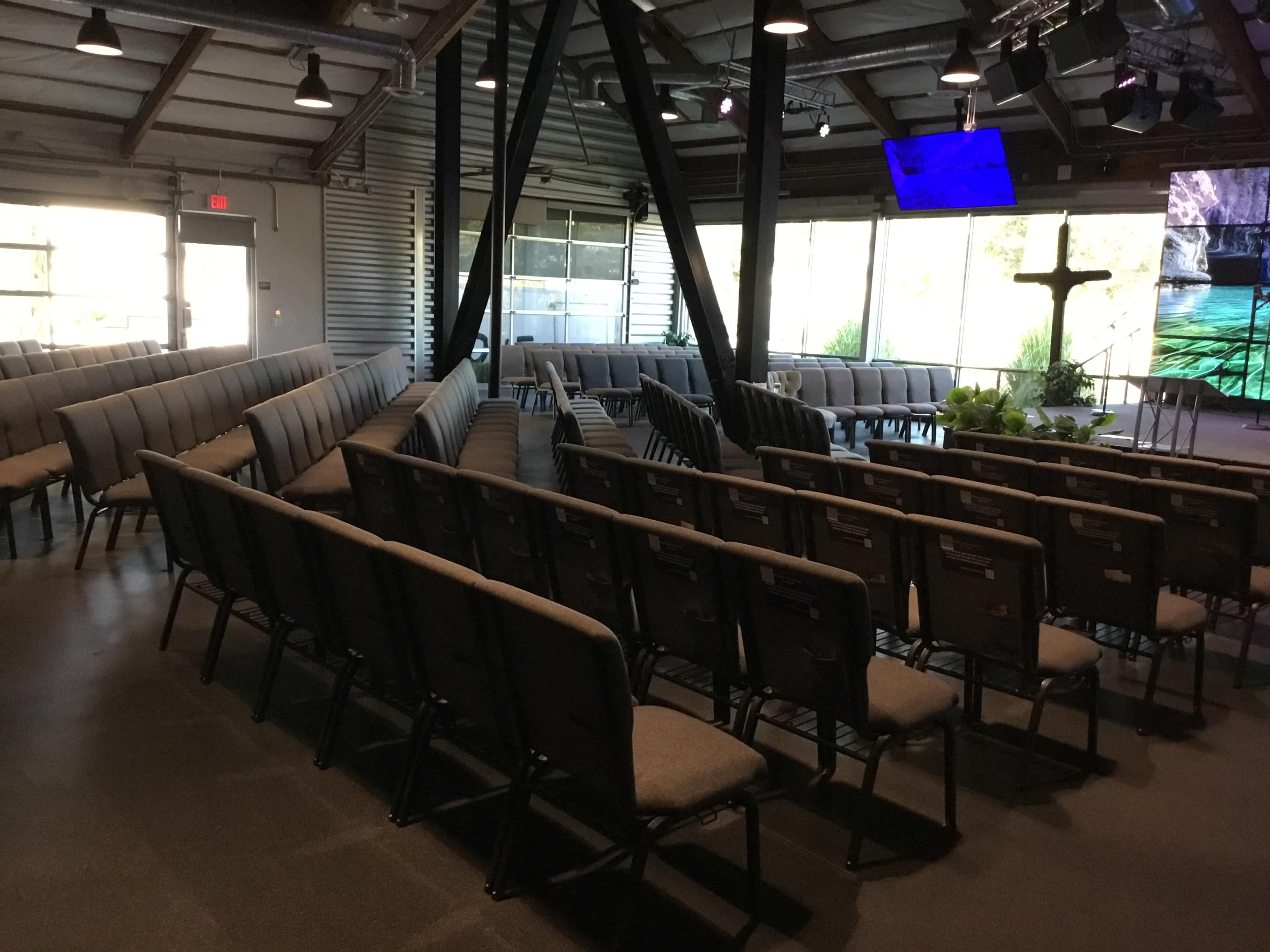 The image shows a spacious interior of a church with rows of gray chairs facing a stage area that features a cross and a screen.